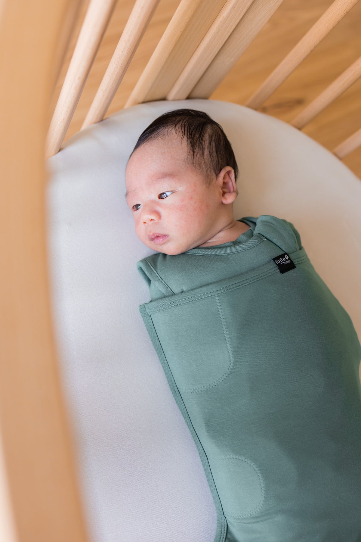 A newborn baby is swaddled in a sage green baby sleep bag swaddler inside a light wood crib.