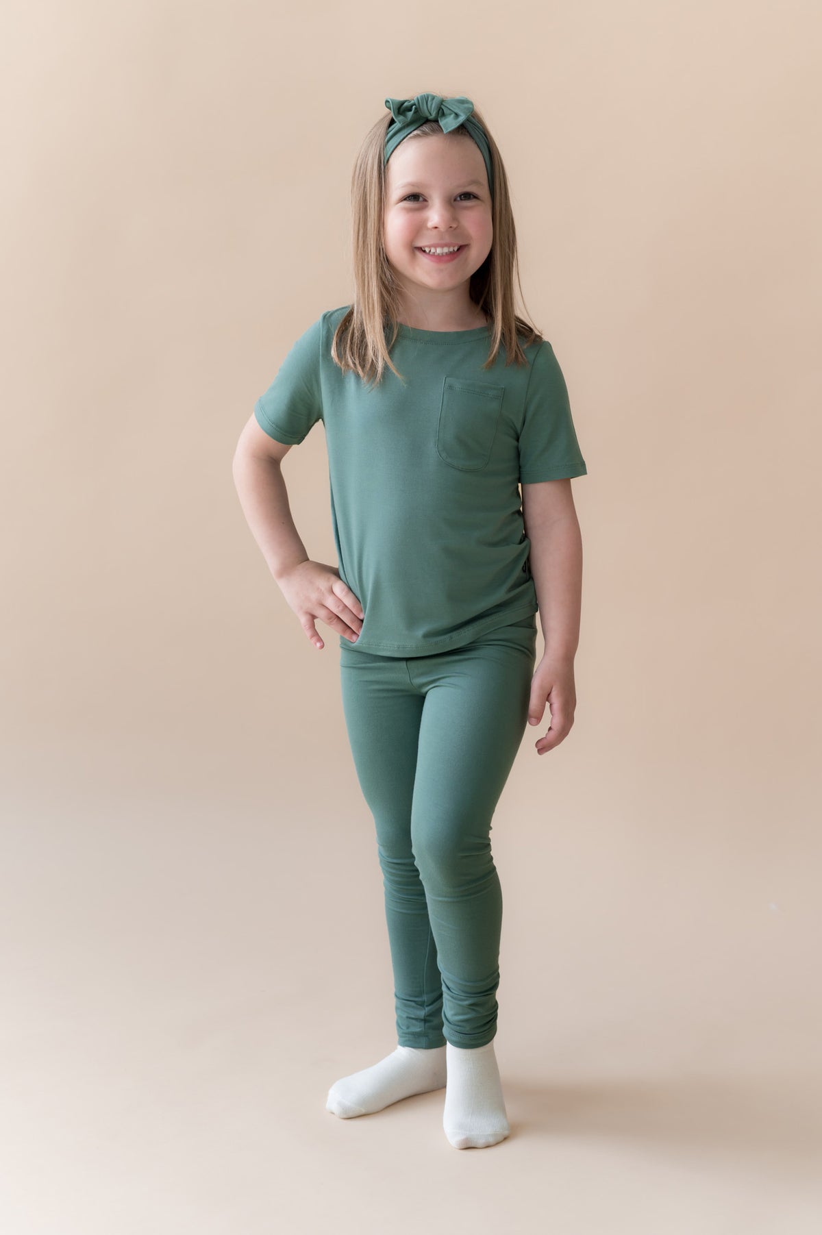 Girl wearing Toddler Leggings in Pine under pine shirt with knotted bow headband in pine on a beige background