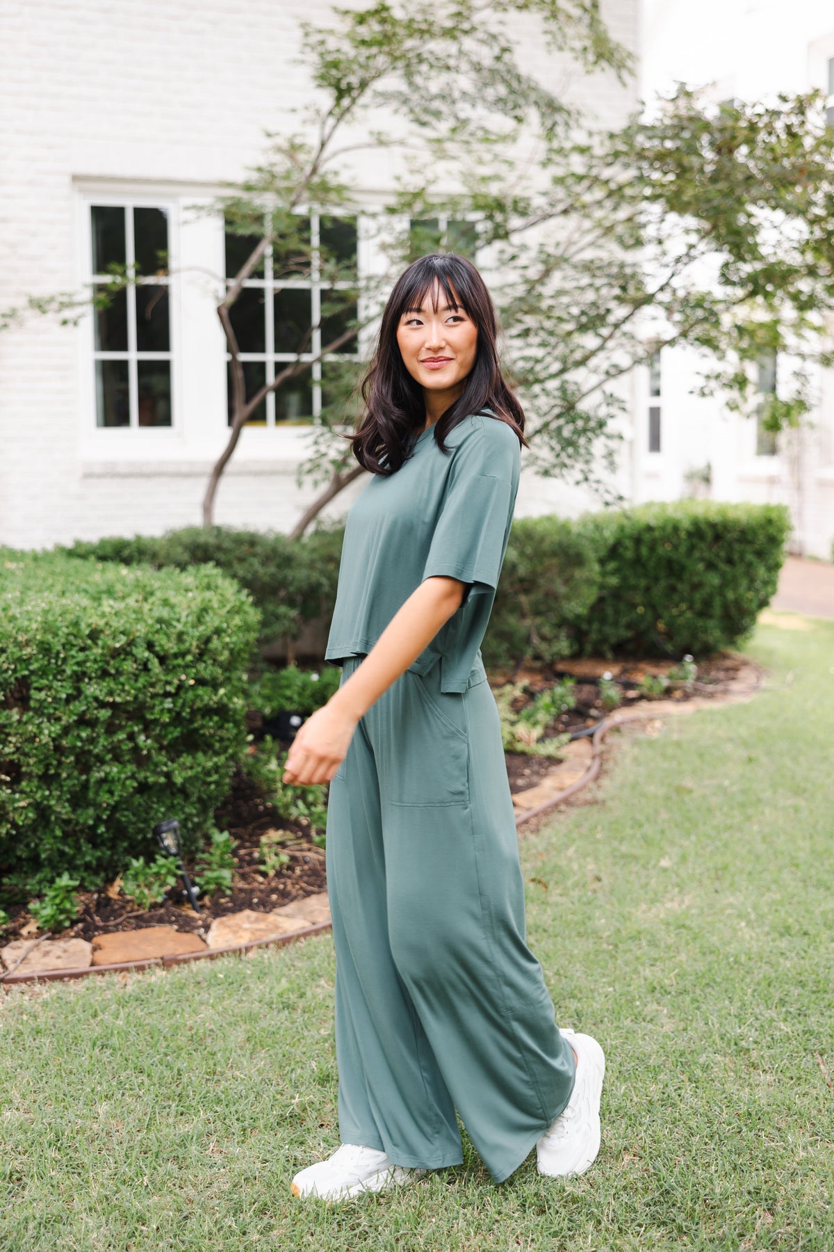 Female model standing outdoors in a garden wearing the Women's Wide Leg Pant in Pine and matching short sleeve tee on white shoes