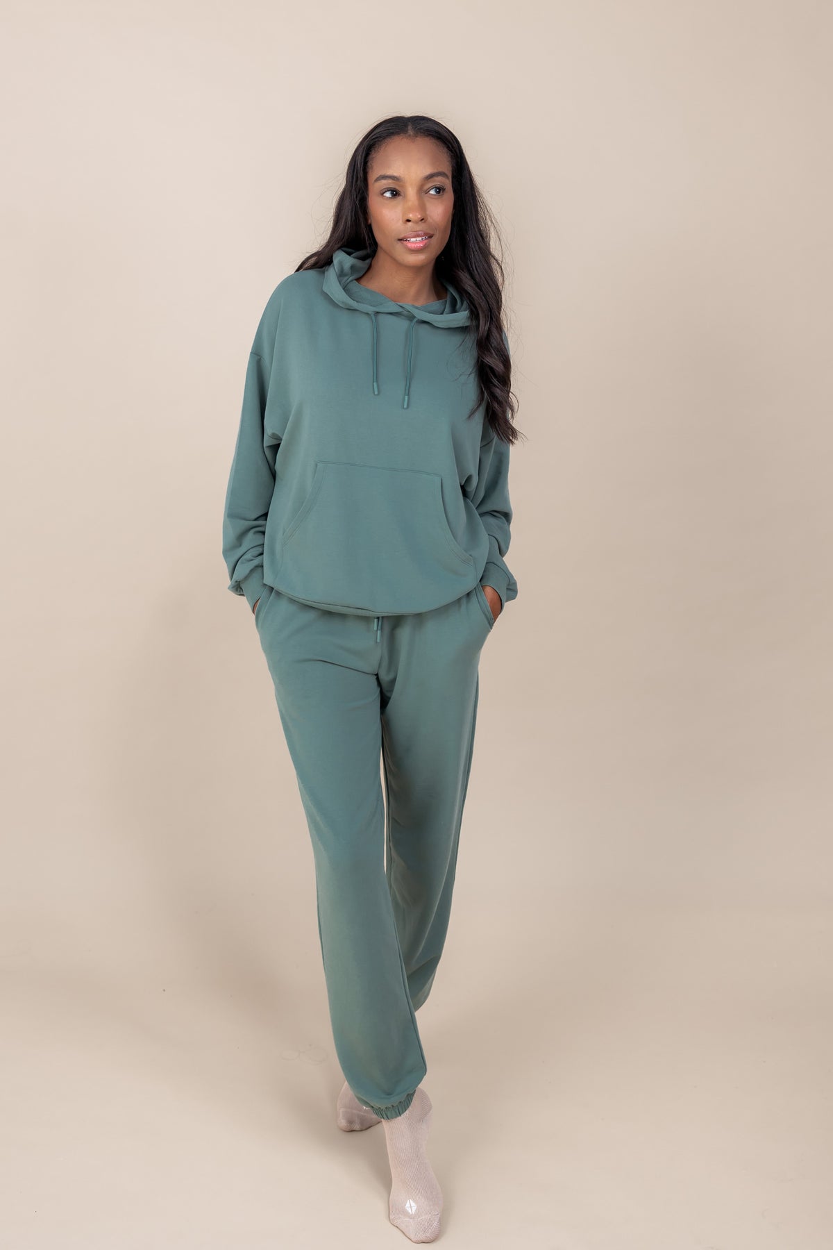 Woman wearing Women's Hoodie in Pine color over a similar color jogger pants on a beige background