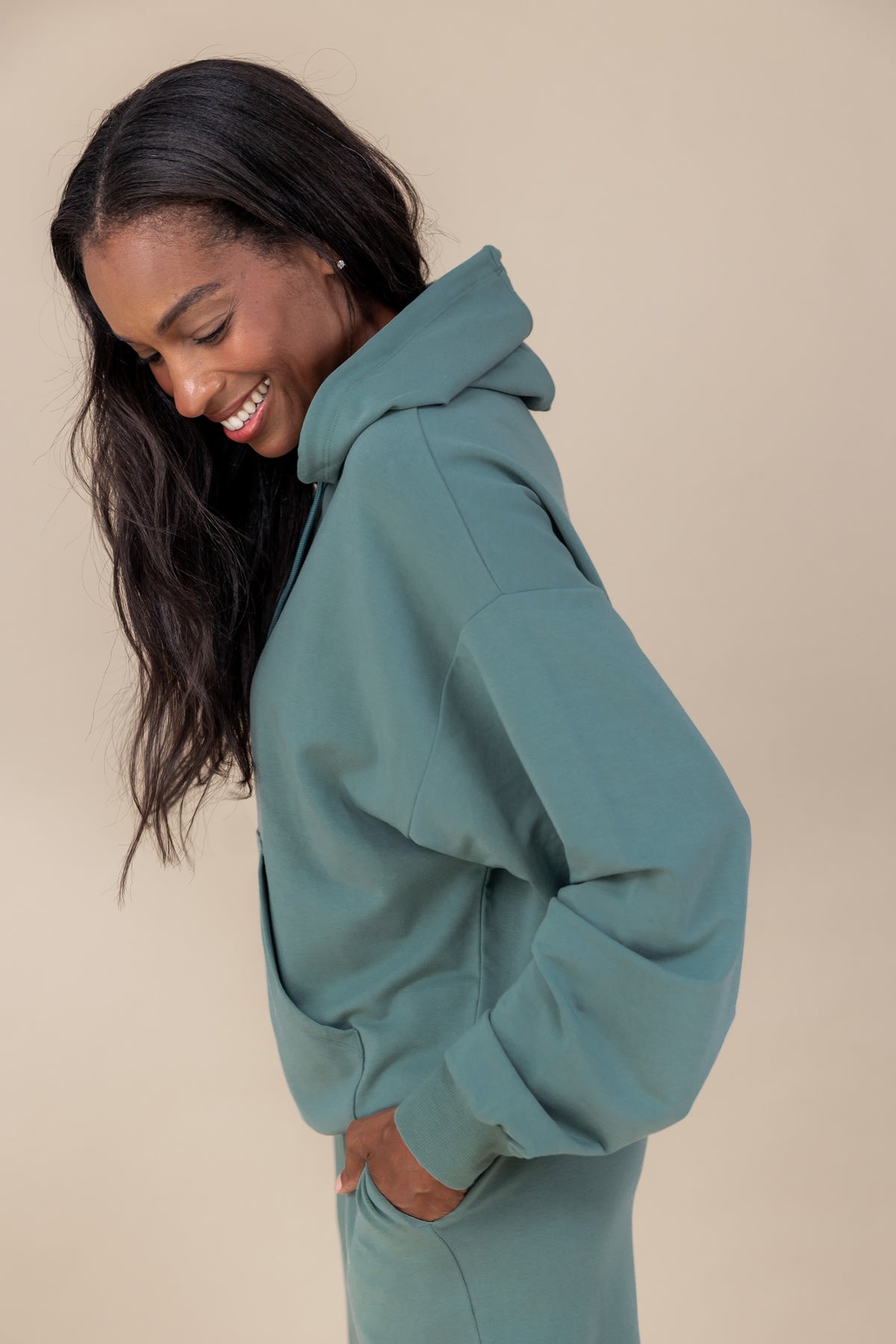 Side pose of Woman wearing Women's Hoodie in Pine color over a similar color jogger pants on a beige background