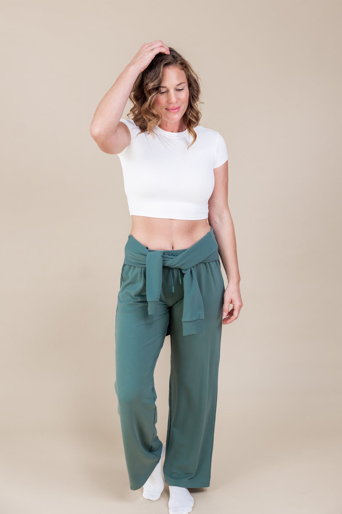 Woman posing in white T-shirt with Women's Sweatshirt in Pine color tied on her waist and Women's sweatpants in pine on a beige background