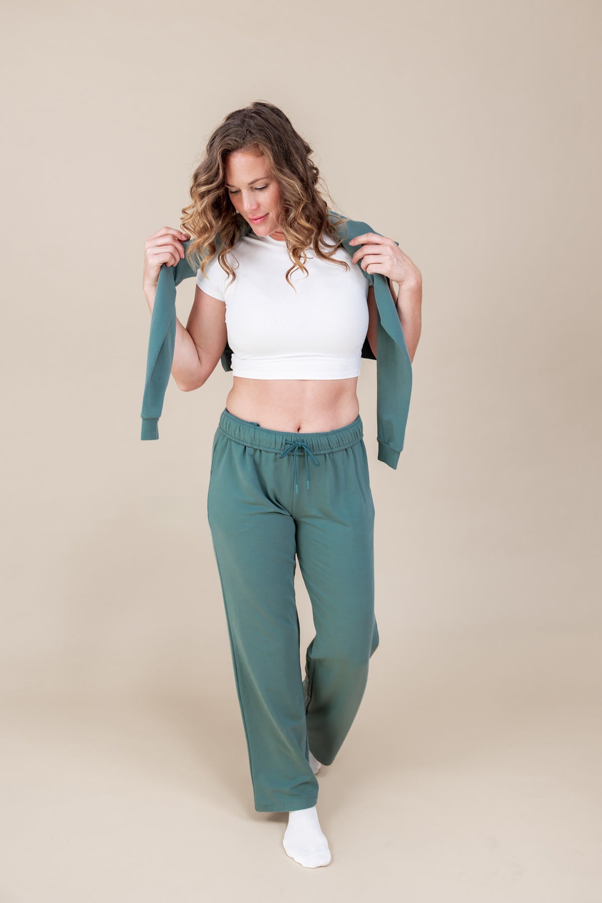 Woman wearing white T-shirt with Women's Sweatshirt in Pine color over her shoulders with Women's sweatpants in pine on a beige background