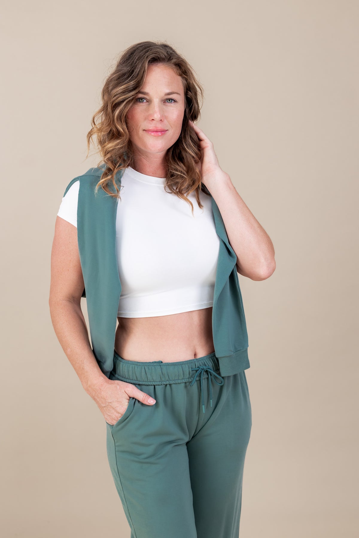 Woman wearing white T-shirt with Women's Sweatshirt in Pine color over her shoulders with Women's sweatpants in pine on a beige background