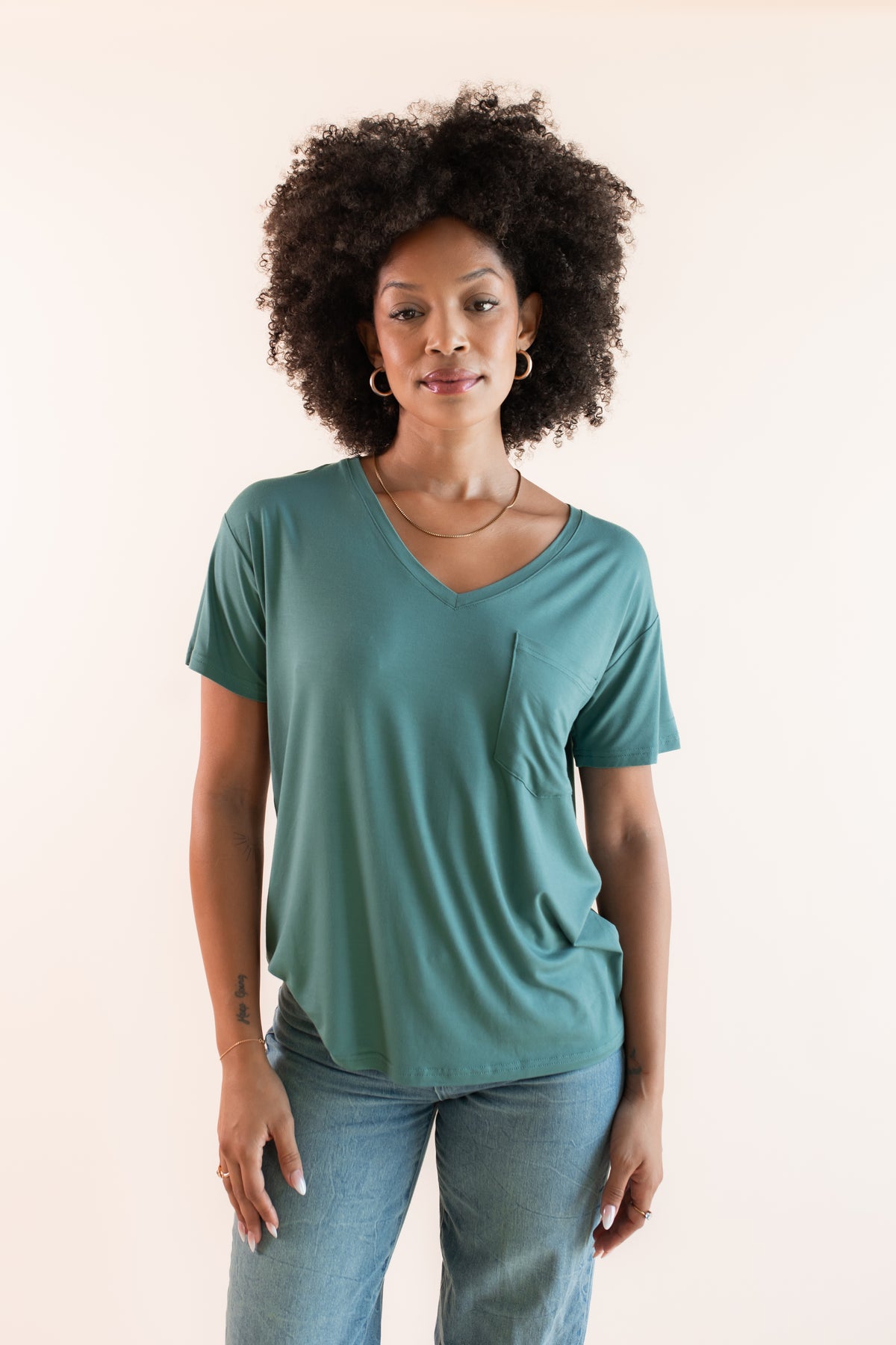 Model posing in Women's Oversized V-Neck Tee in Pine over blue jeans on a beige background