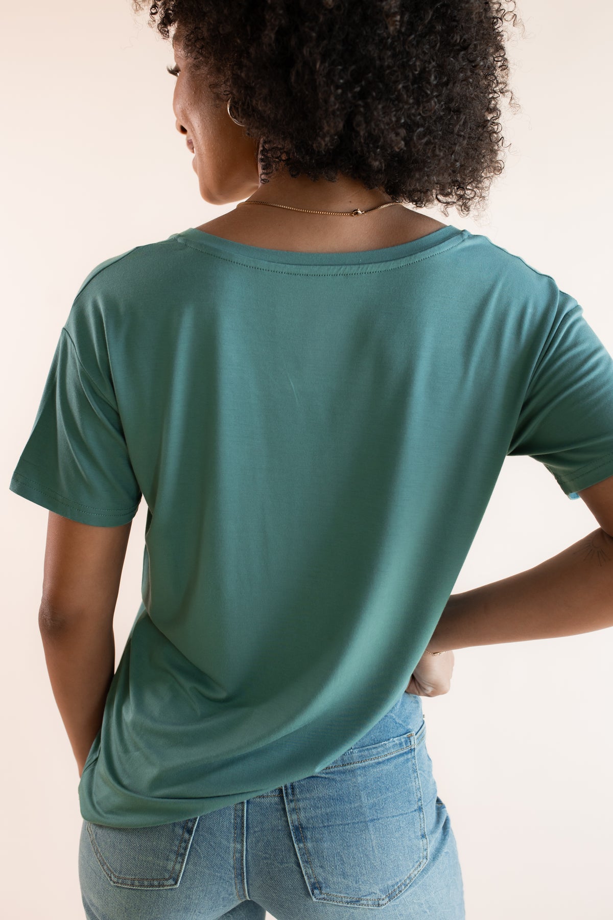 Model showcasing back side of Women's Oversized V-Neck Tee in Pine over blue jeans on a beige background