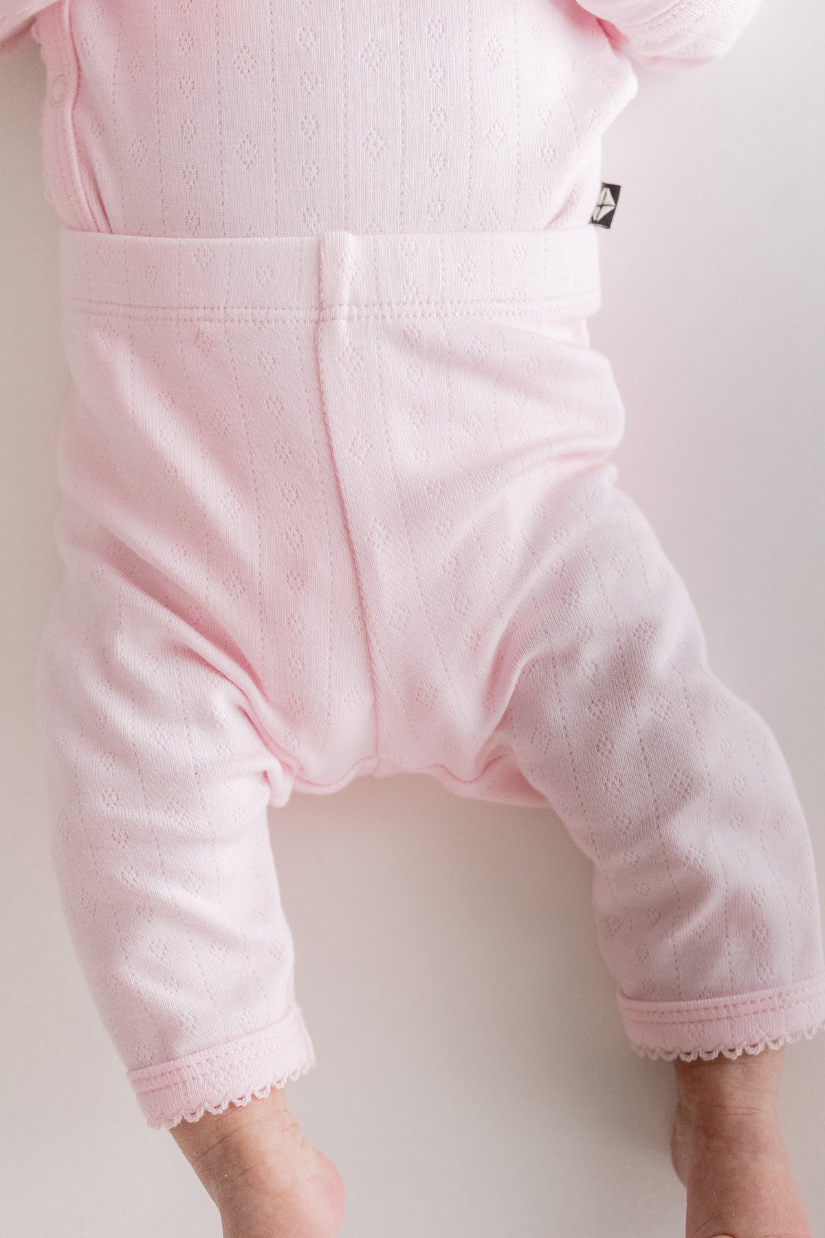 Close-up of a baby wearing light pink pointelle pants with a wide waistband and scalloped hem, paired with a matching pink top.