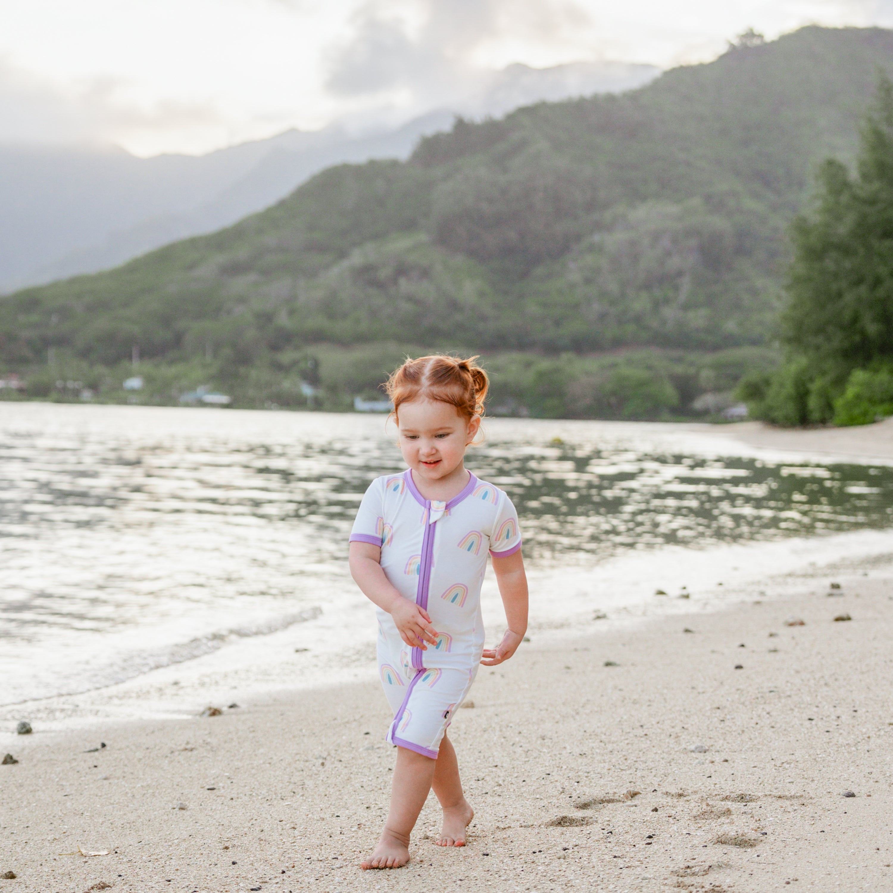 Toddler walking on the beach wearing bamboo Kyte Baby Zippered Shortall Romper in Poi Rainbow