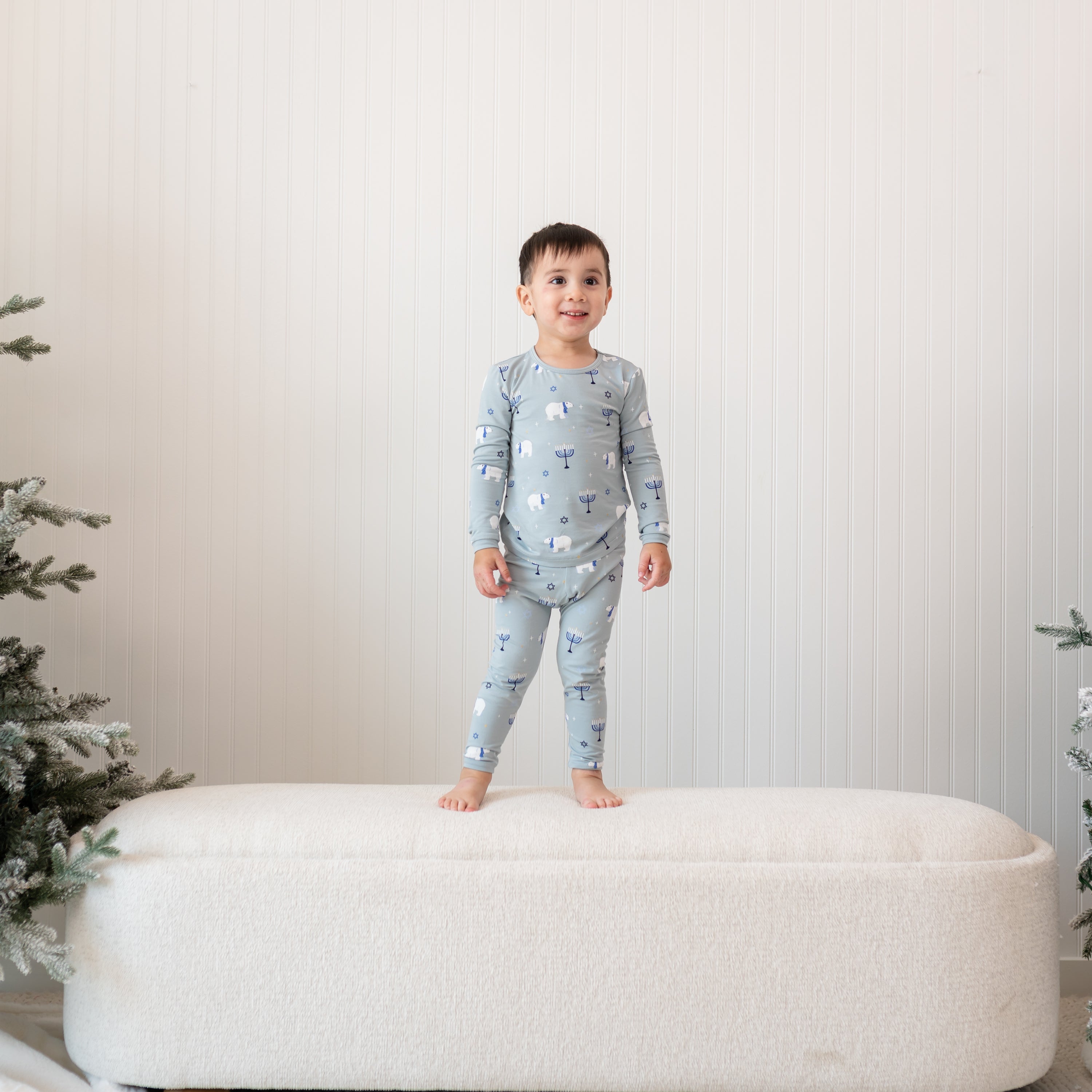 Young boy standing on a cream ottoman in front of a white paneled wall wearing the Long Sleeve Pajamas in Polar Lights