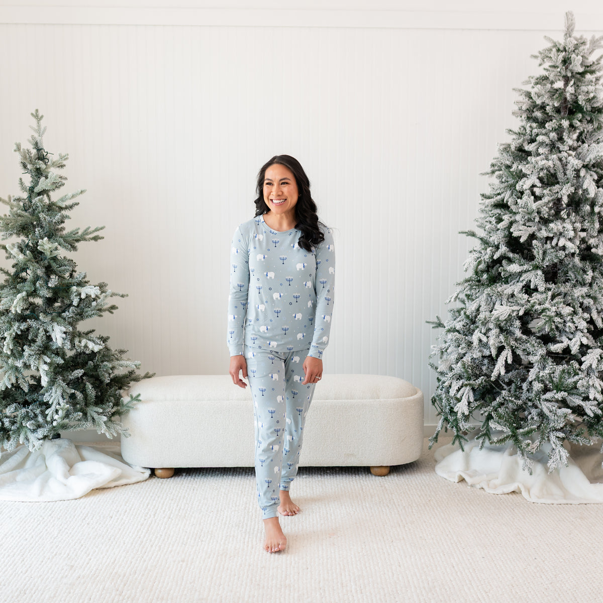 Smiling female model wearing the Women's Jogger Pajama Set in Polar Lights standing between two frosted trees in front of a white paneled wall and cream ottoman