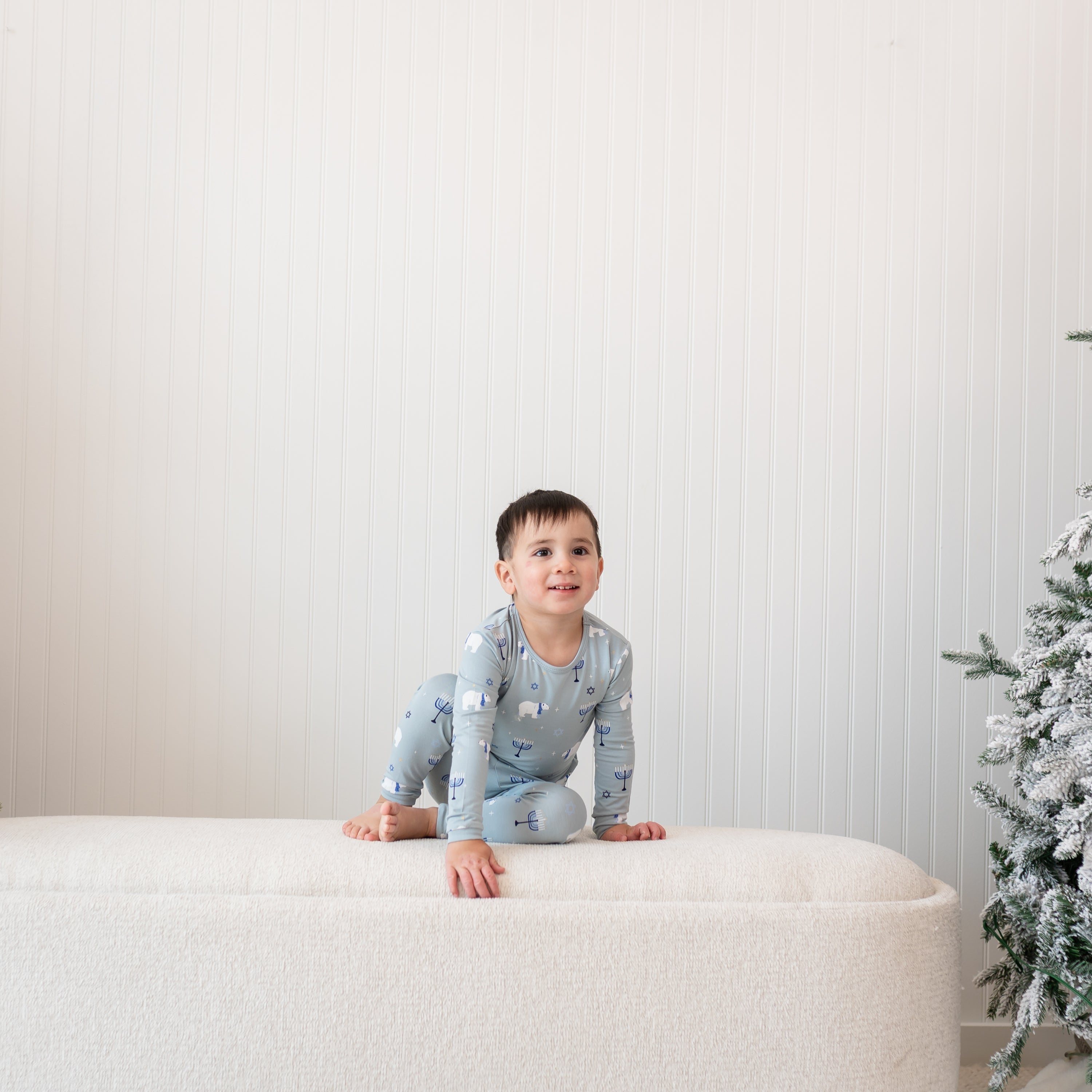 Young boy sitting on a cream ottoman wearing the Long Sleeve Pajamas in Polar Lights