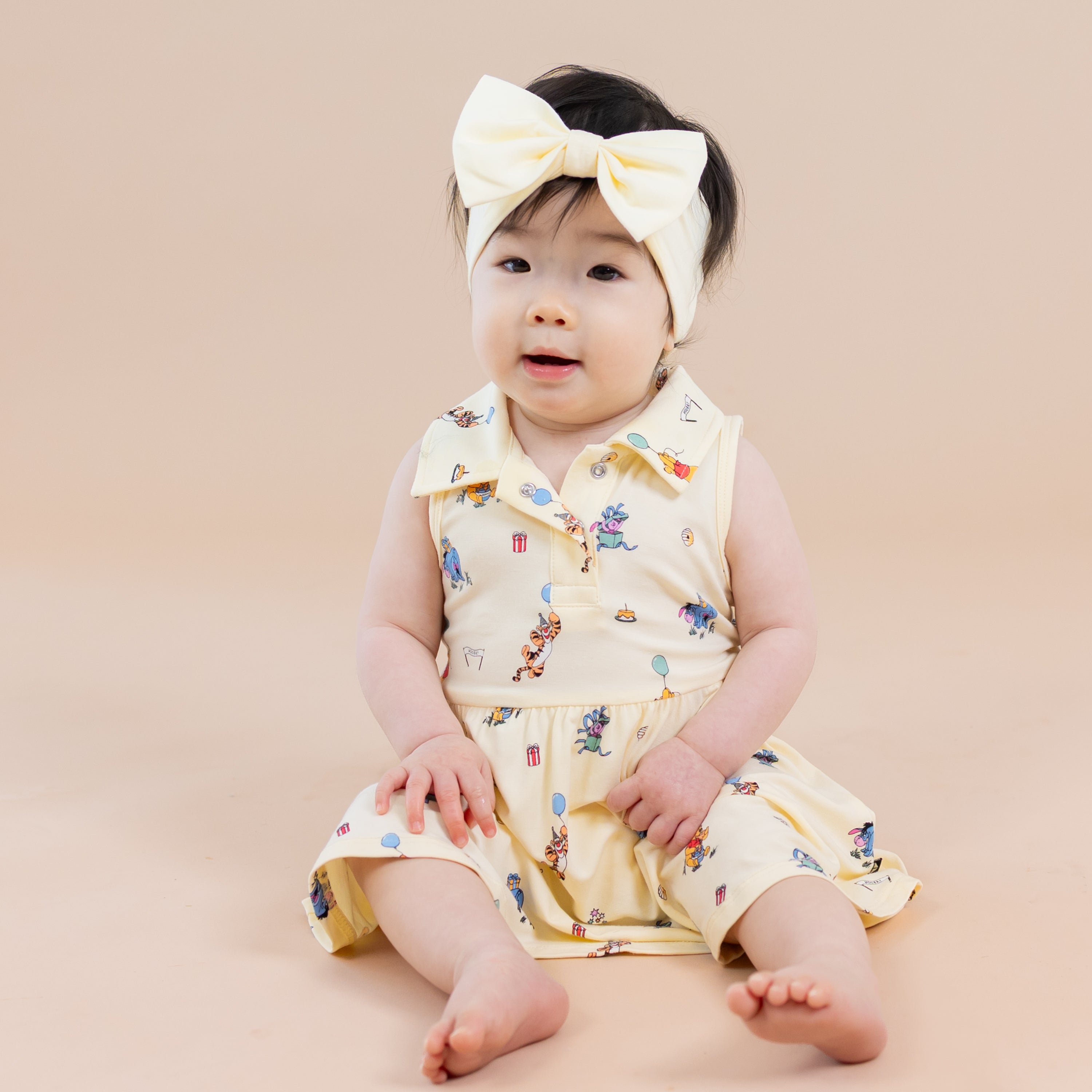 Infant sitting on the floor wearing the Polo Bodysuit Dress in Winnie the Pooh Celebration with a light yellow bow