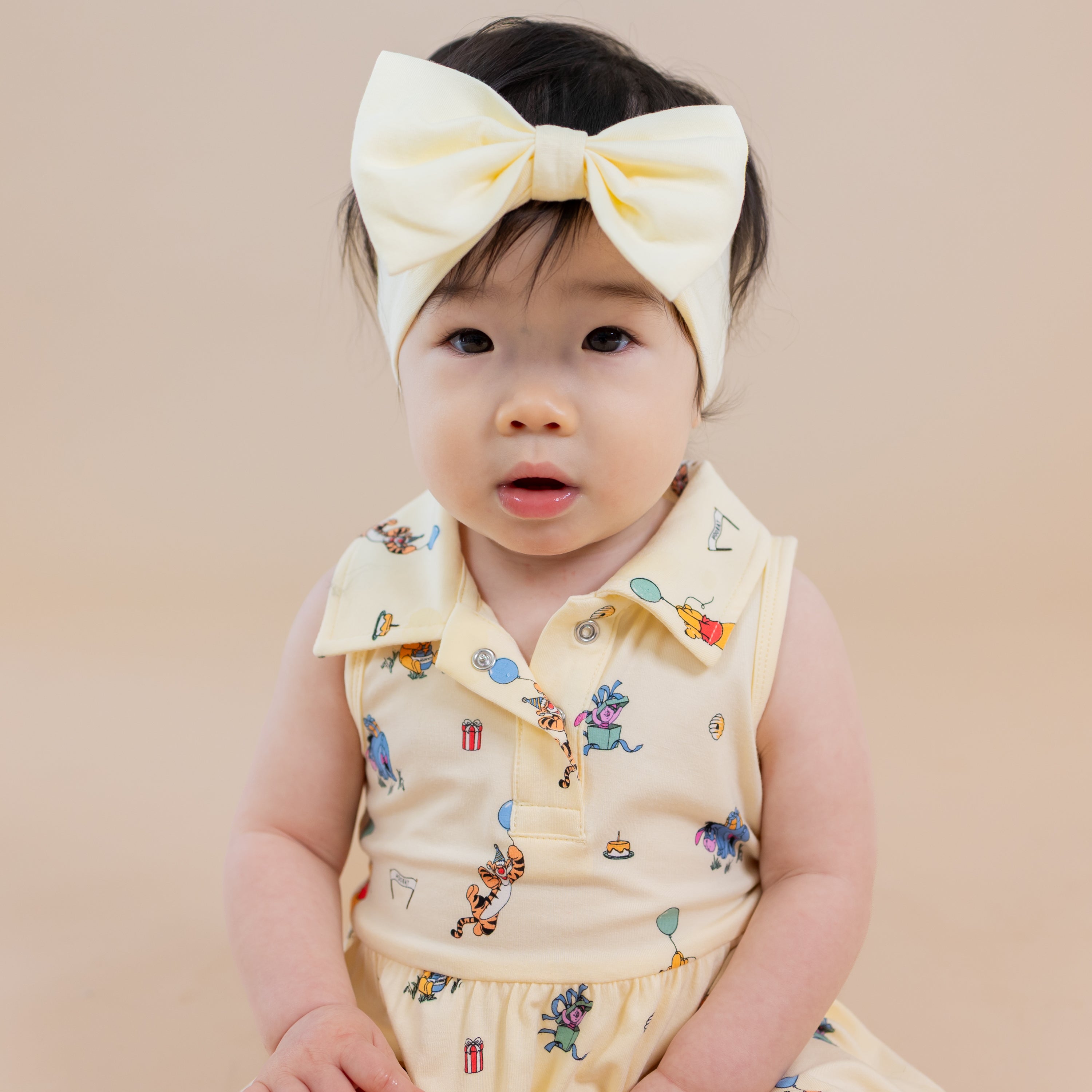 Close up of infant sitting wearing the Polo Bodysuit Dress in Winnie the Pooh Celebration with light yellow bow