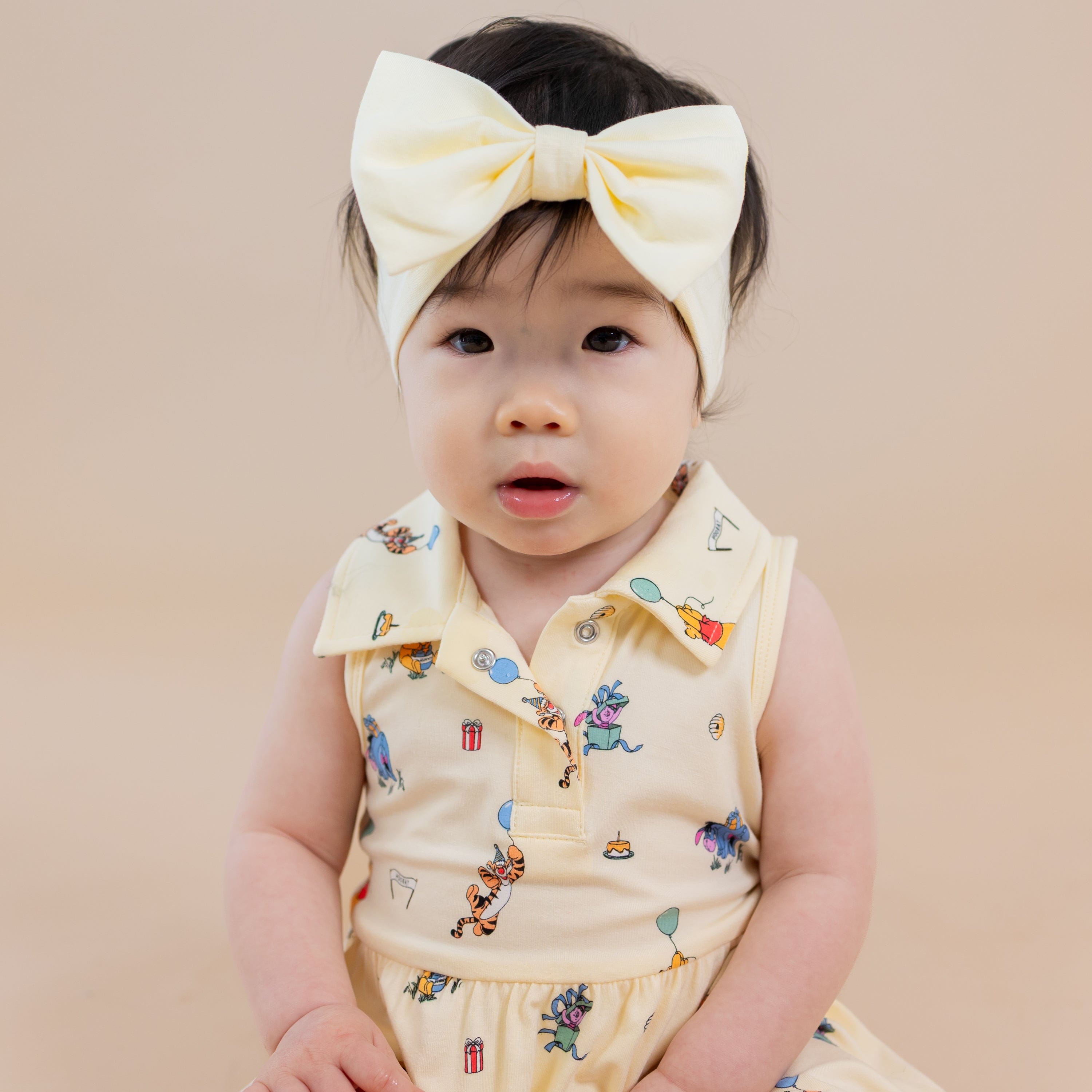 Close up of infant sitting wearing the Polo Bodysuit Dress in Winnie the Pooh Celebration with light yellow bow
