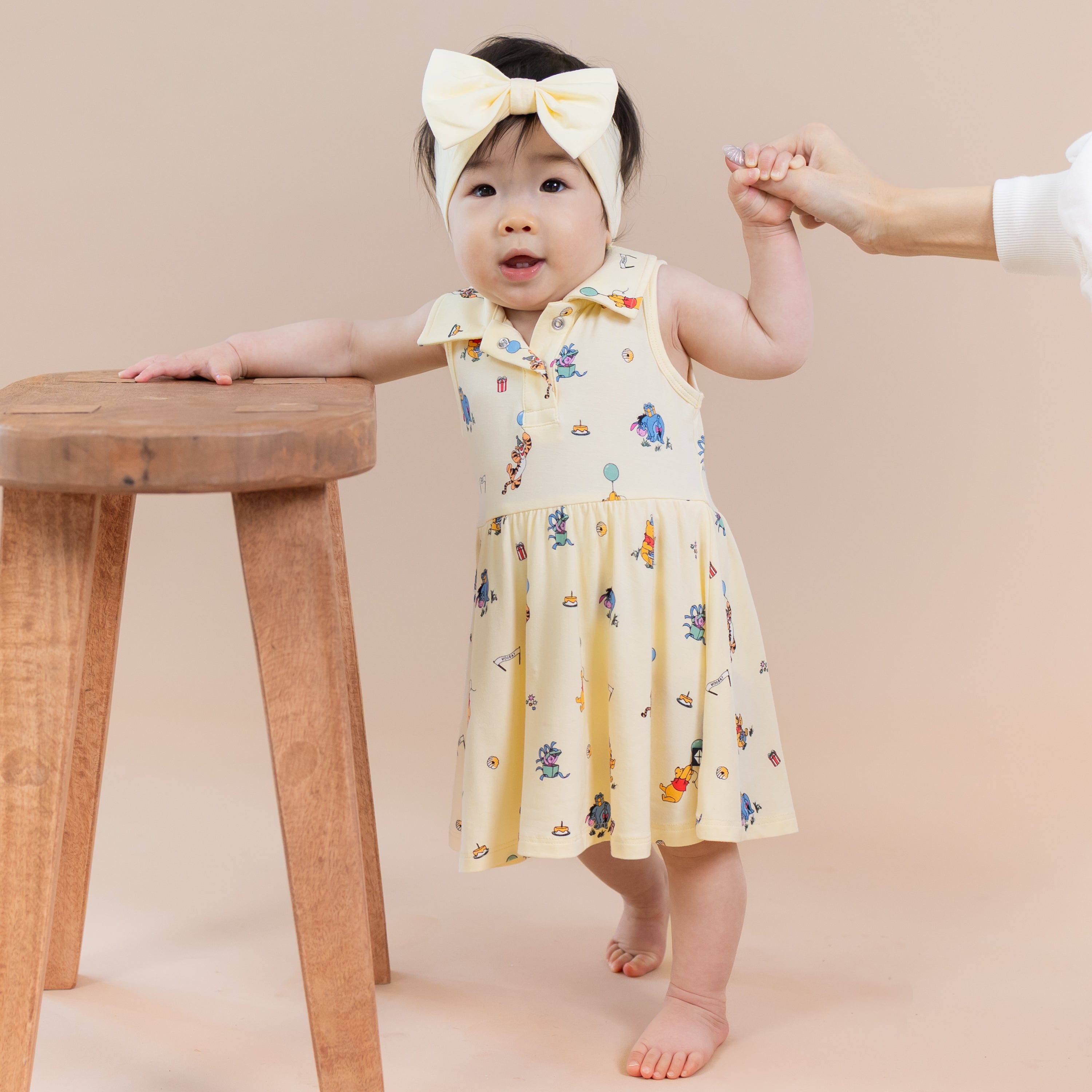 Infant standing beside a wooden stool wearing the Polo Bodysuit Dress in Winnie the Pooh Celebration holding on to her mothers hand