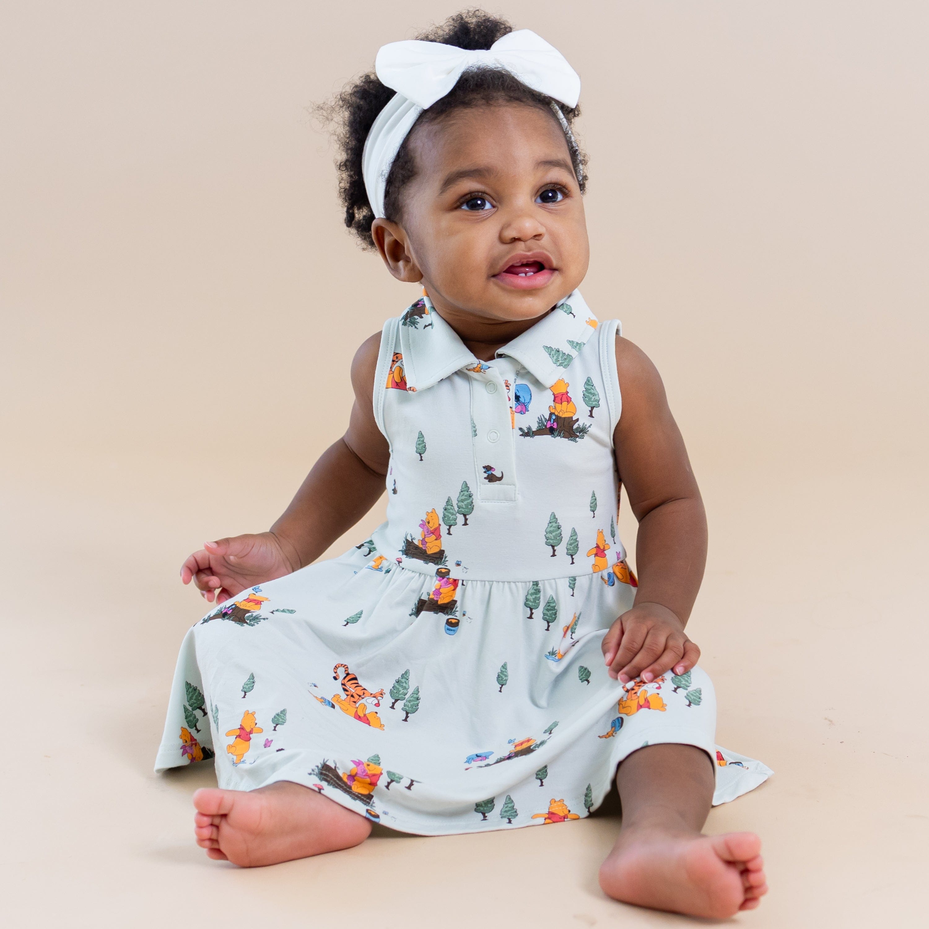 Young toddler sitting on the floor wearing the Polo Bodysuit Dress in Winnie the Pooh Hundred Acre Wood paired with a white bow