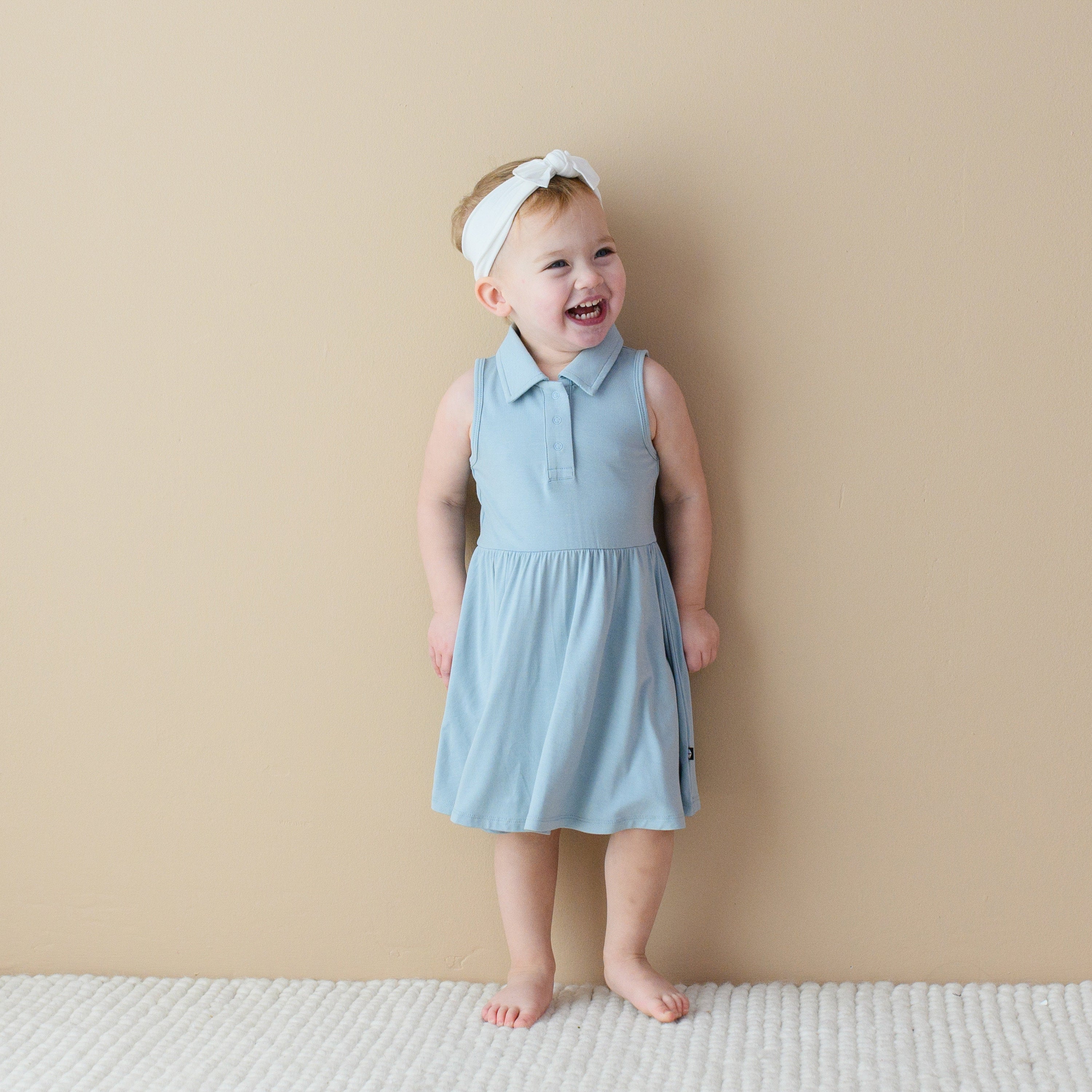 Toddler standing in-front of a tan wall while wearing a Polo Bodysuit Dress in Fog