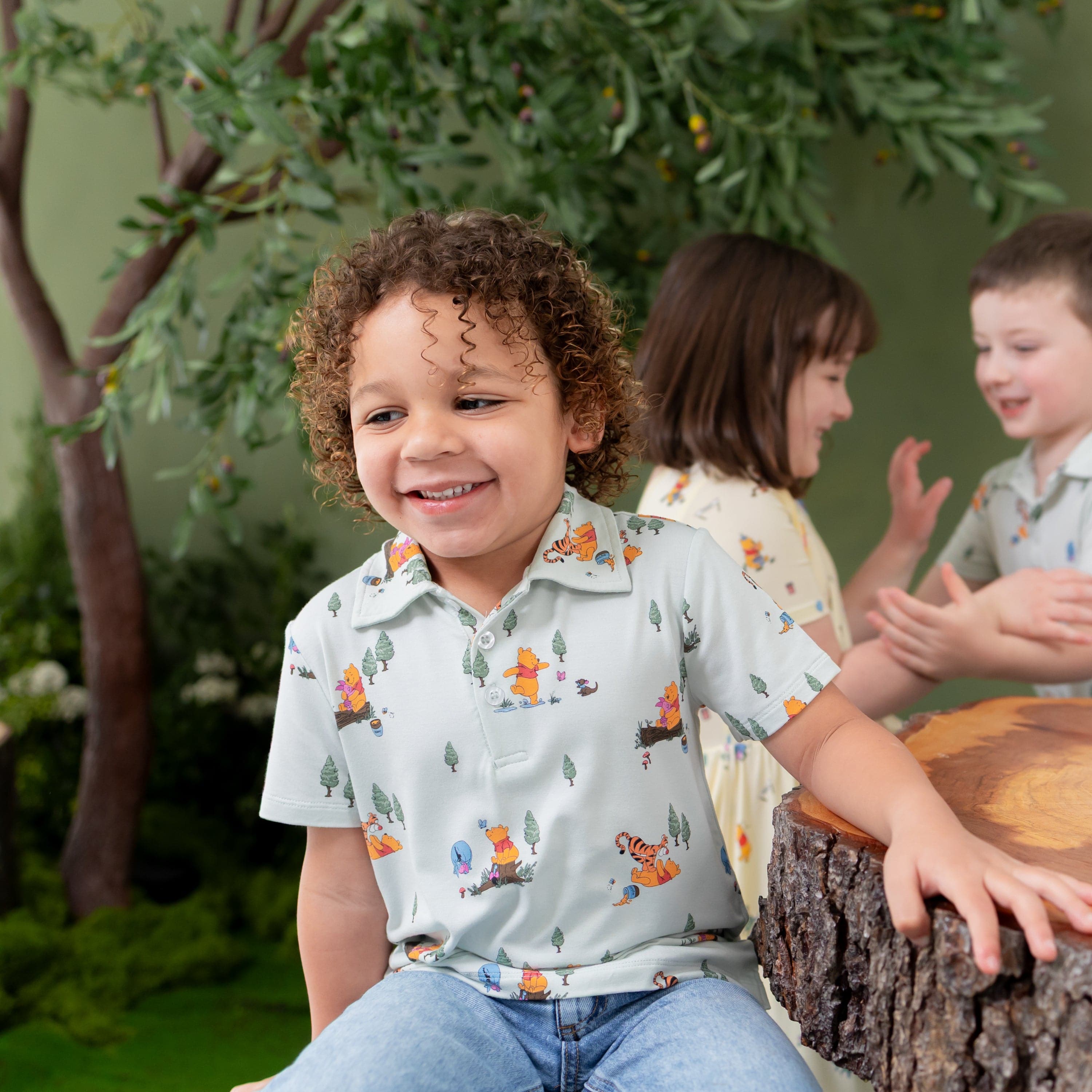 Young boy sitting beside a tree stumpe wearing the Toddler Short Sleeve Polo in Winnie the Pooh Hundred Acre Wood in front of a tree prop with other children in the background