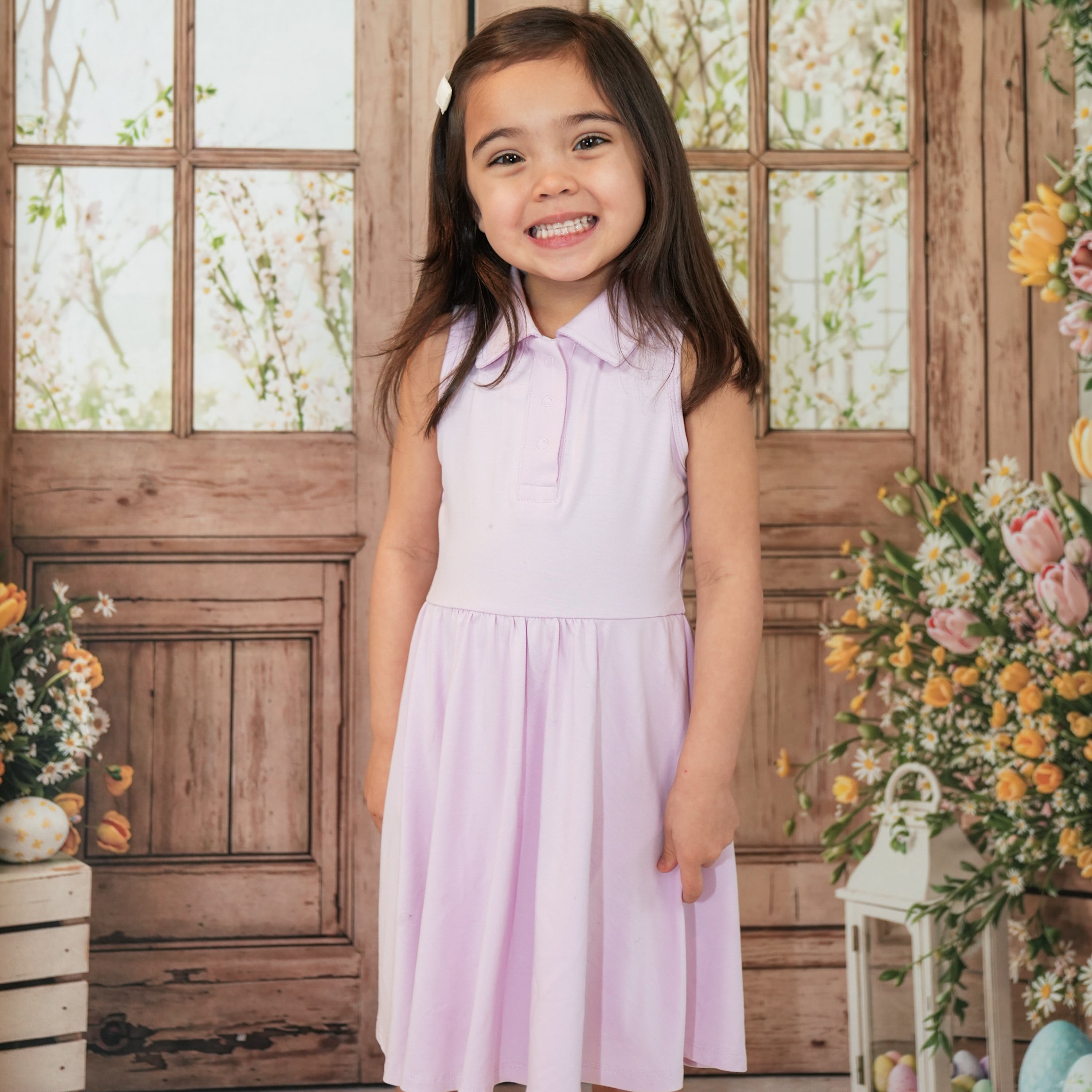 Smiling girl standing in front of a wooden window frame and flowers wearing the Polo Bodysuit Dress in Thistle