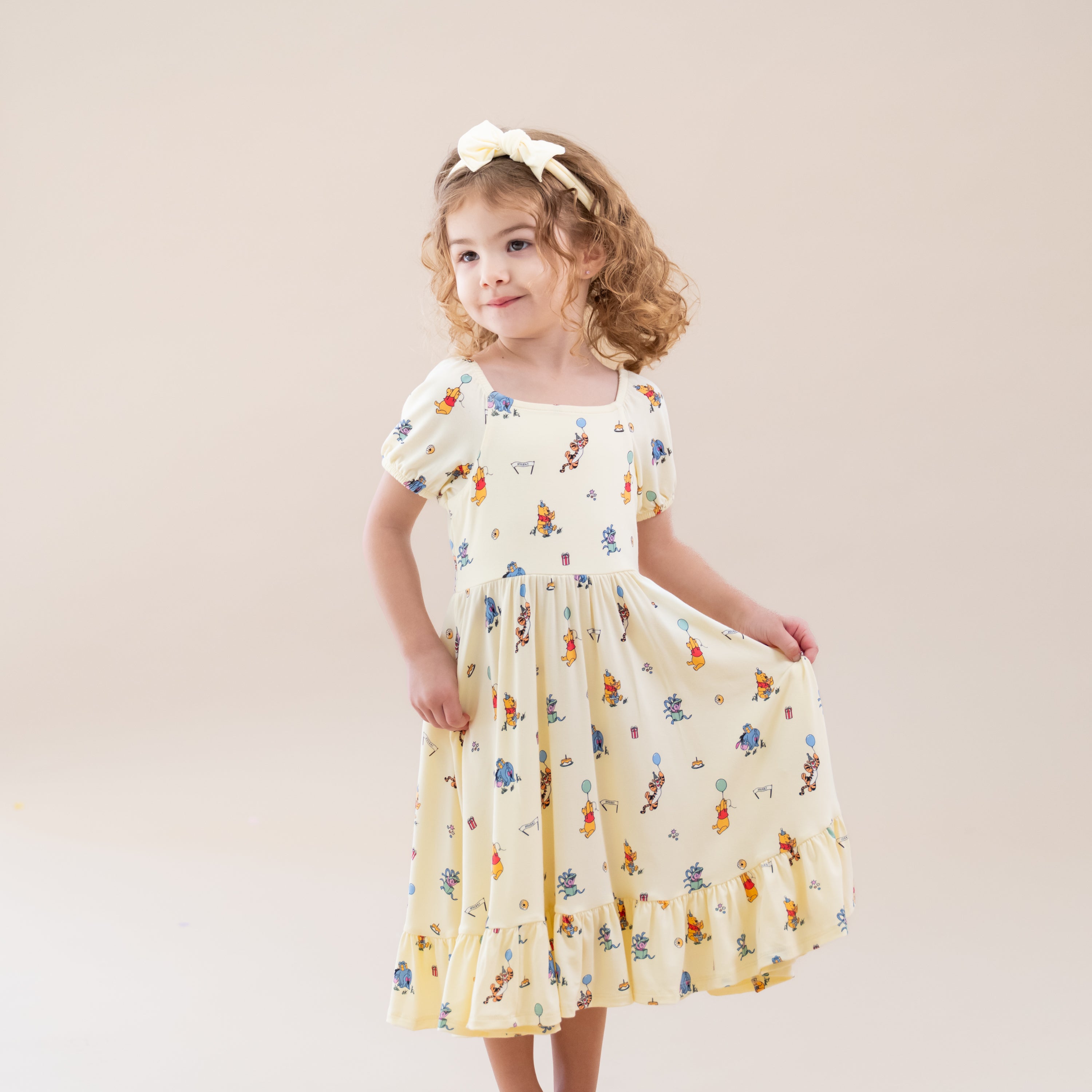 Smiling girl modeling the Puff Sleeve Twirl Dress in Winnie the Pooh Celebration holding the skirt of the dress