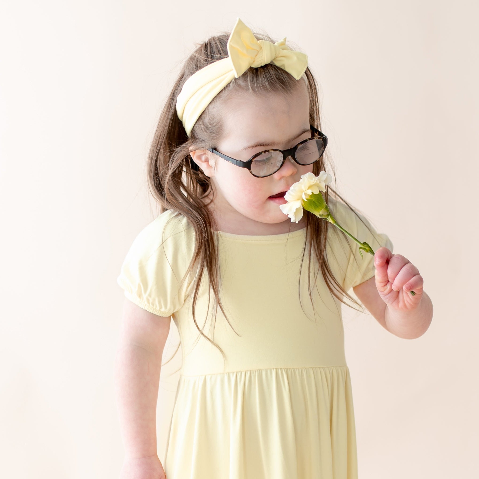 Child wearing a yellow dress, yellow bow and glasses, holding a flower against a plain background