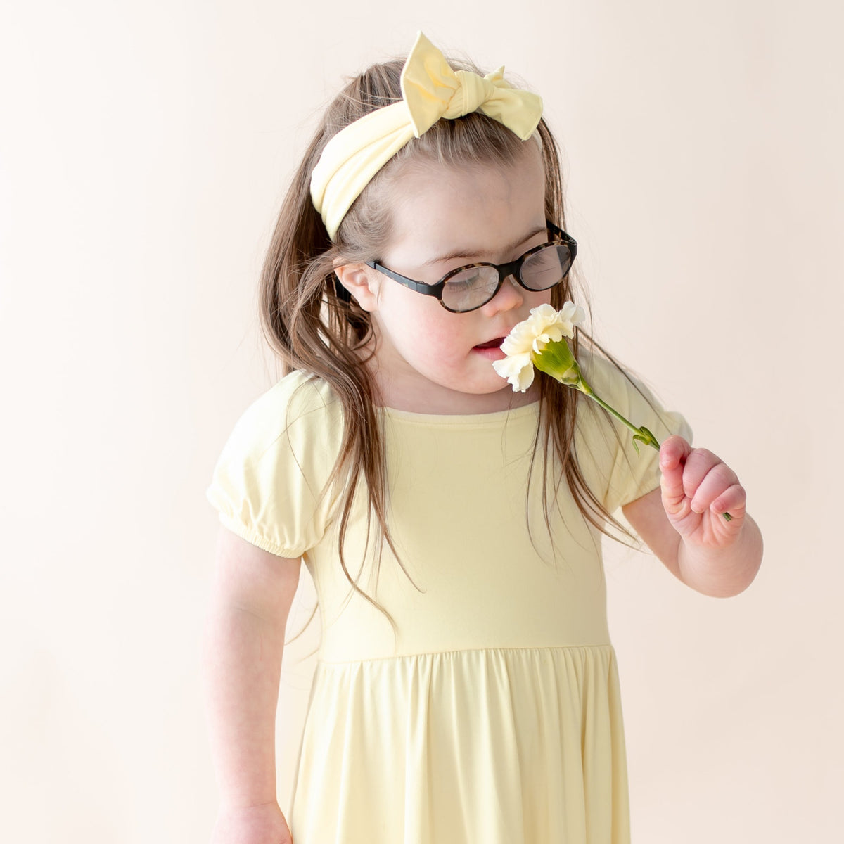 Child wearing a yellow dress, yellow bow and glasses, holding a flower against a plain background