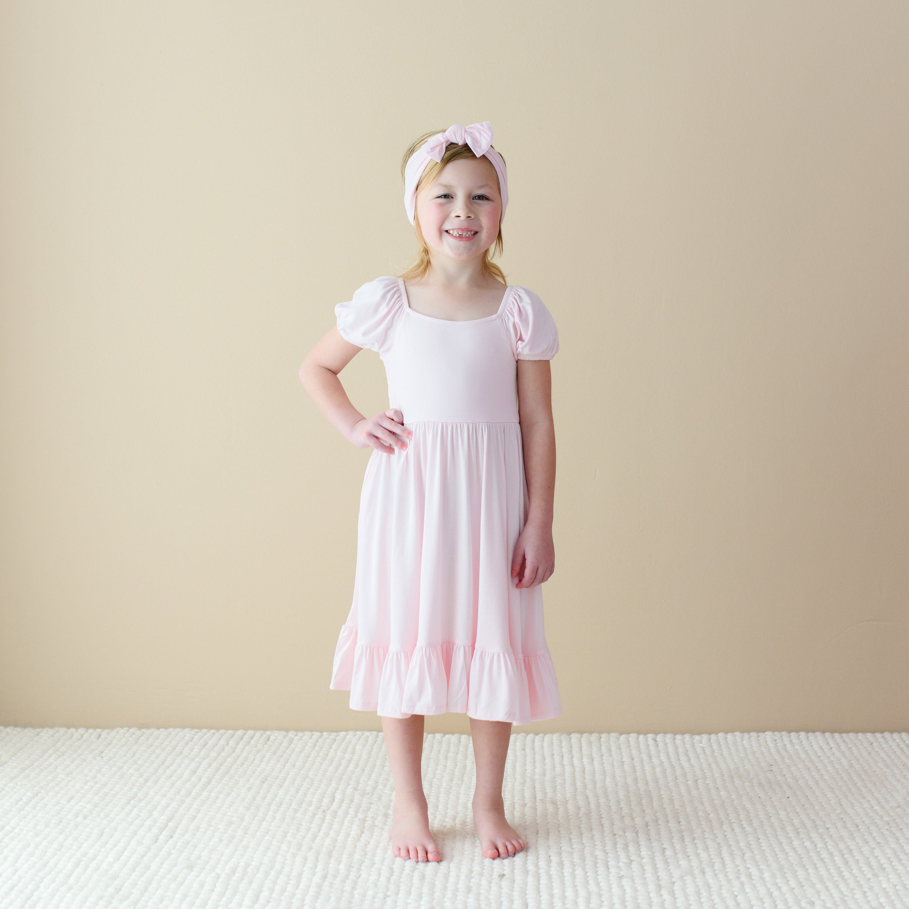 Girl wearing Puff Sleeve Twirl Dress in Sakura Pink with Matching Bow Headband