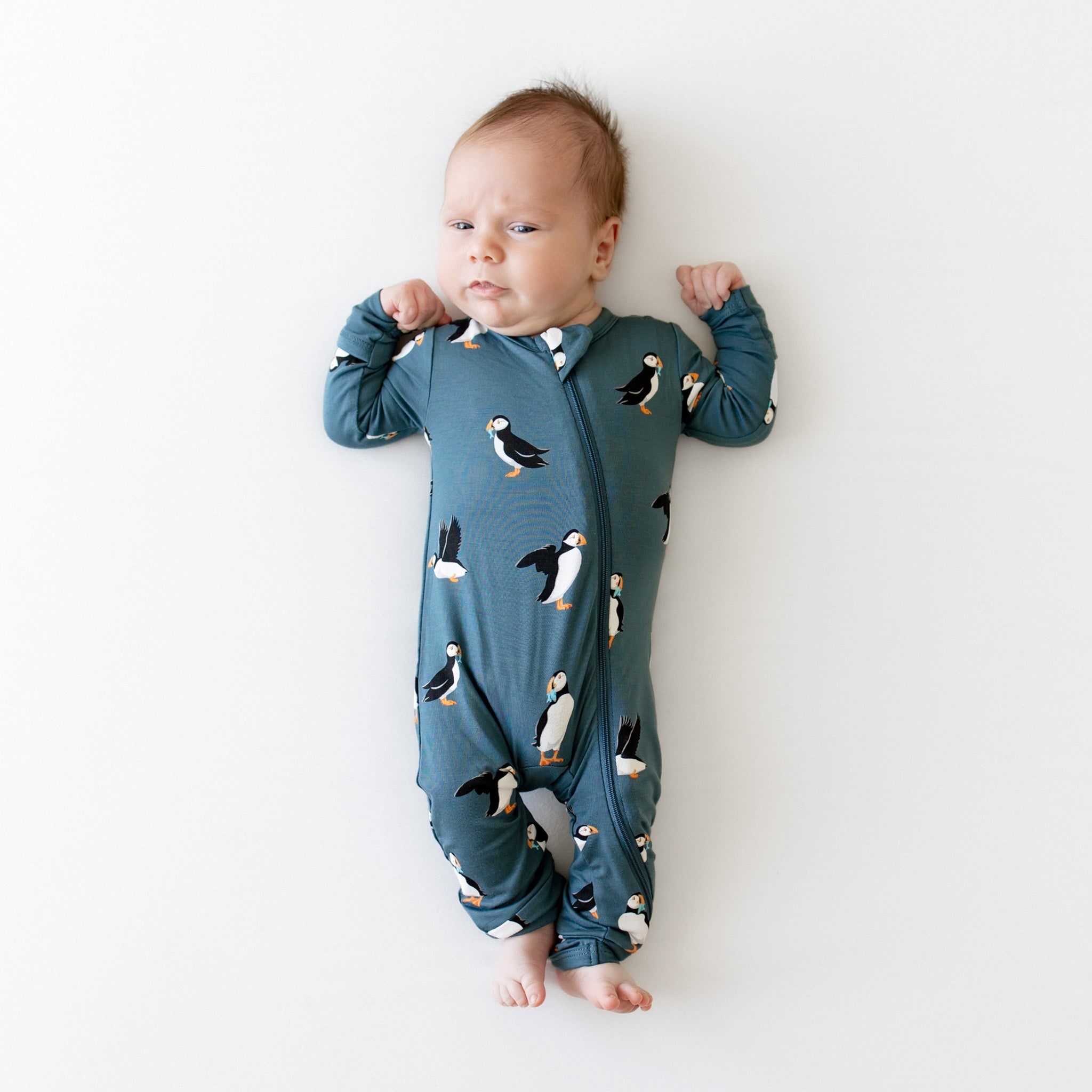 Infant laying on a light neutral surface wearing the Zippered Romper in Puffin