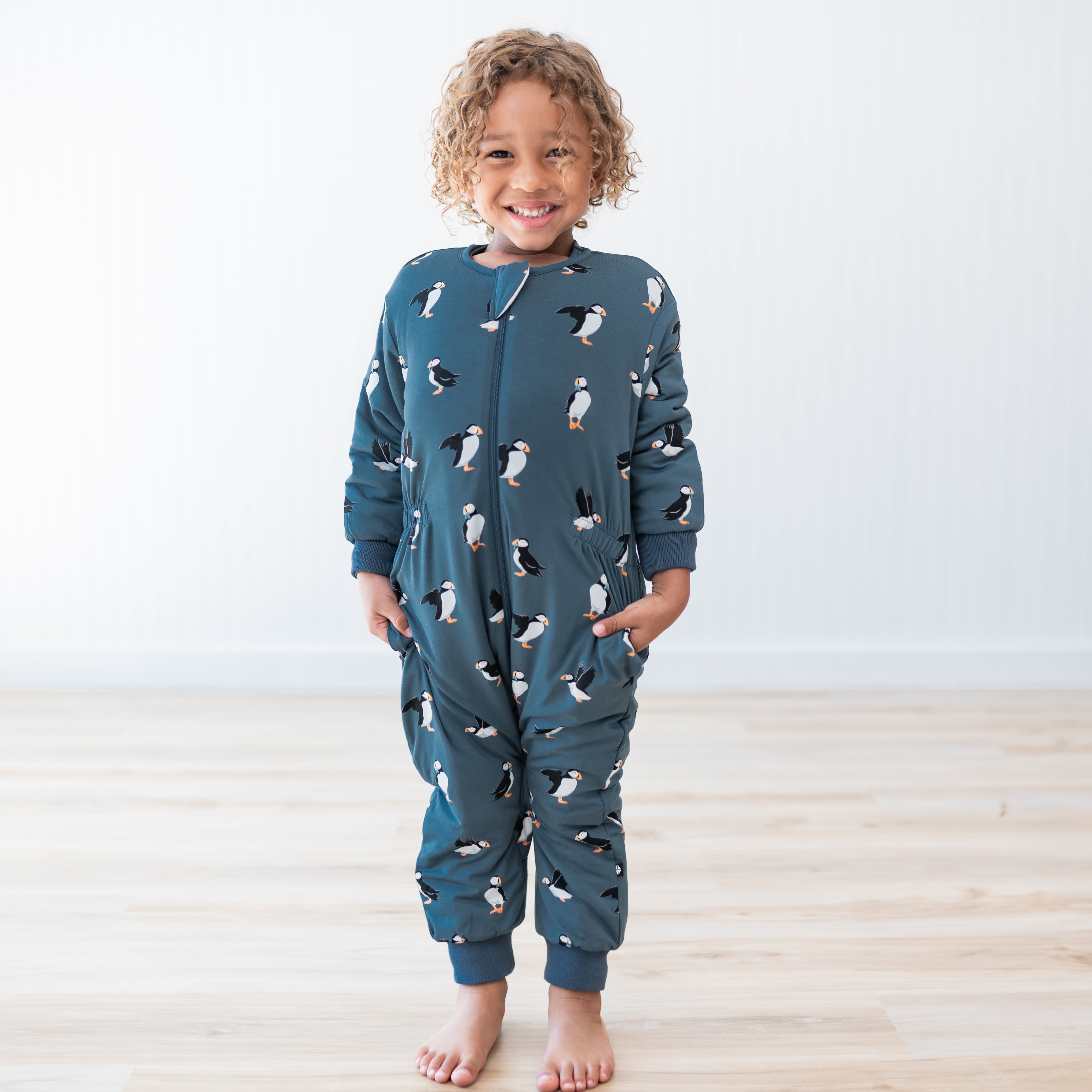 Young model standing straight with hands in the pockets of the Cozy Playsuit in Puffin in front of a white paneled wall