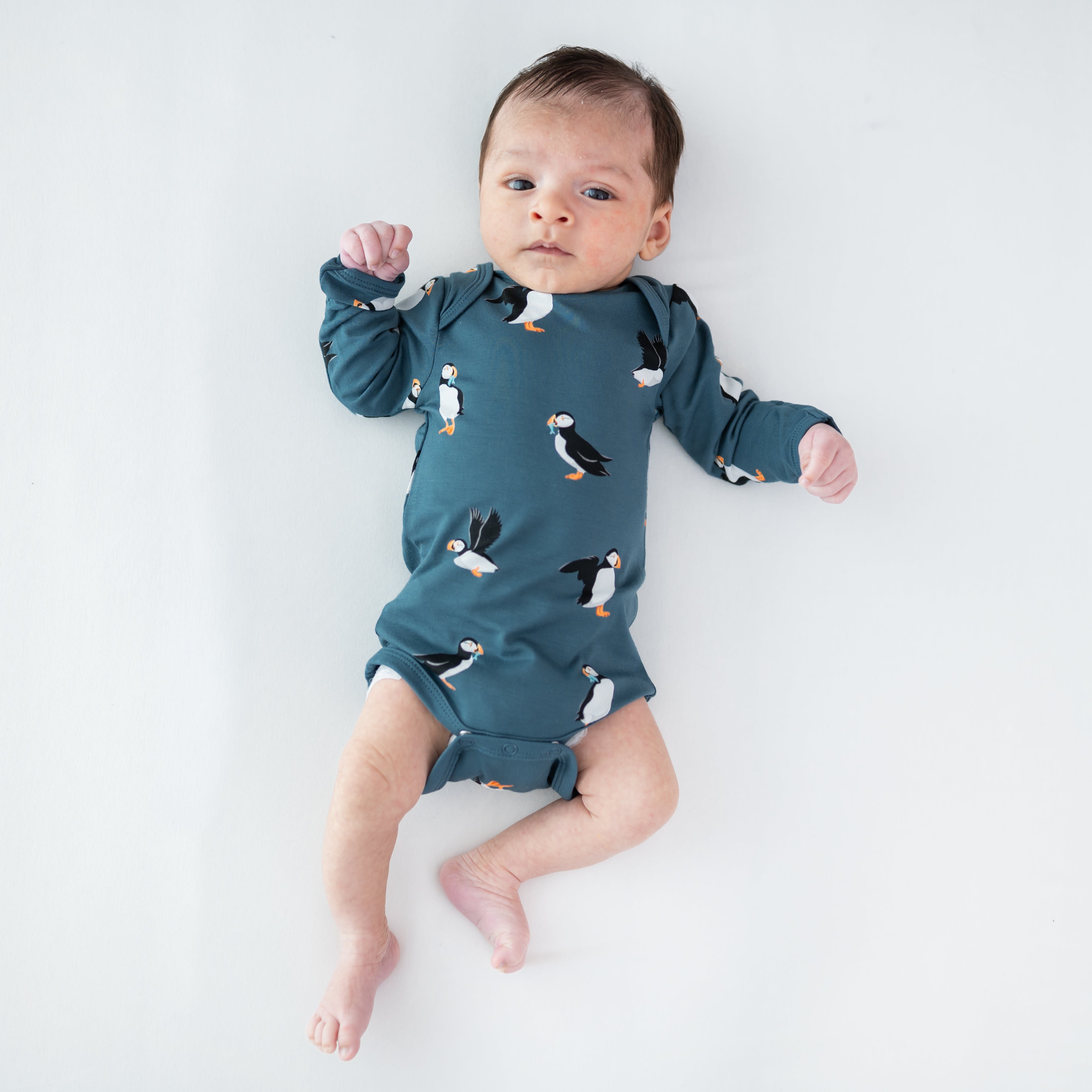 Infant laying on a light neutral surface wearing the Long Sleeve Bodysuit in Puffin
