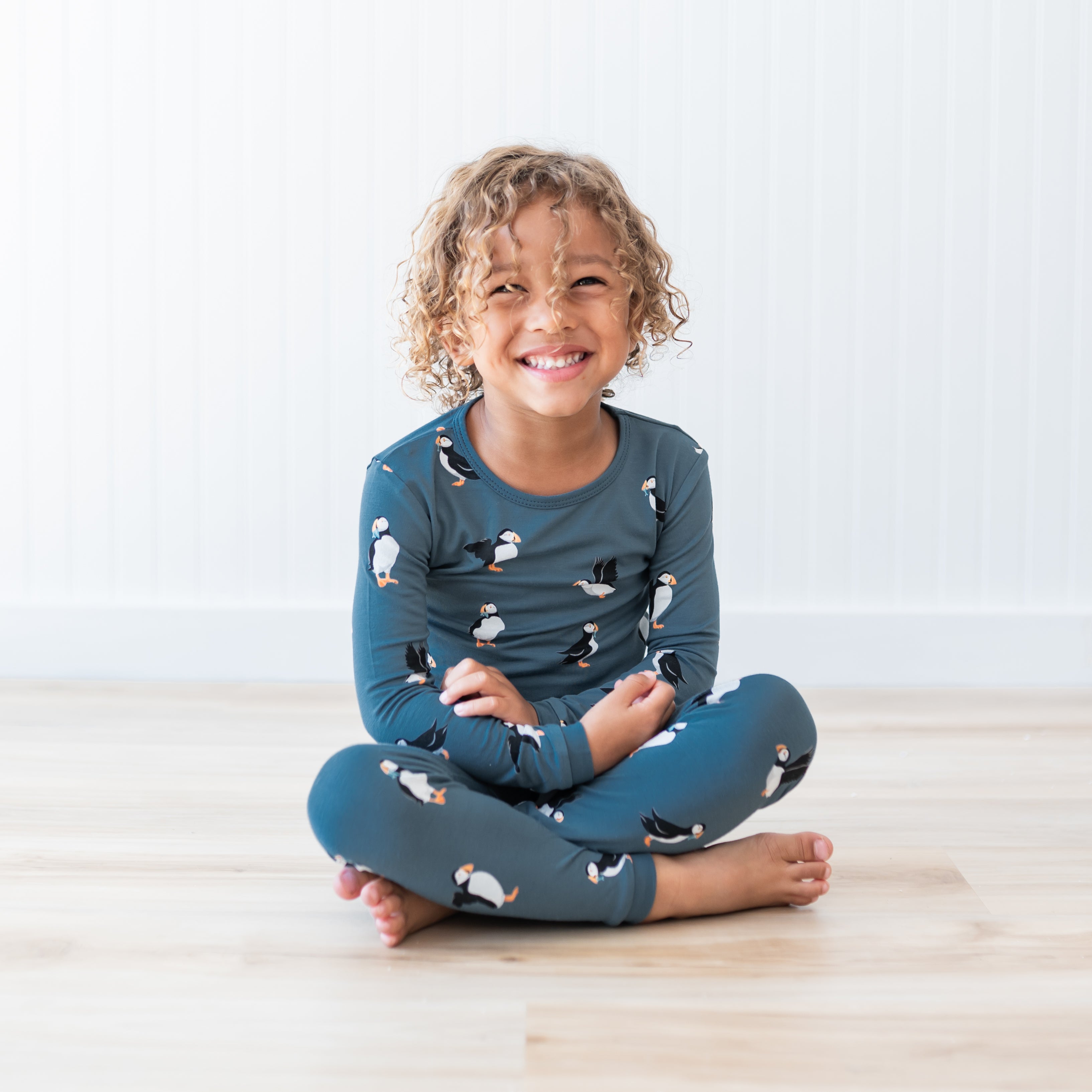 Young smiling child sitting cross legged on a light wood grain floor wearing the Long Sleeve Pajamas in Puffin