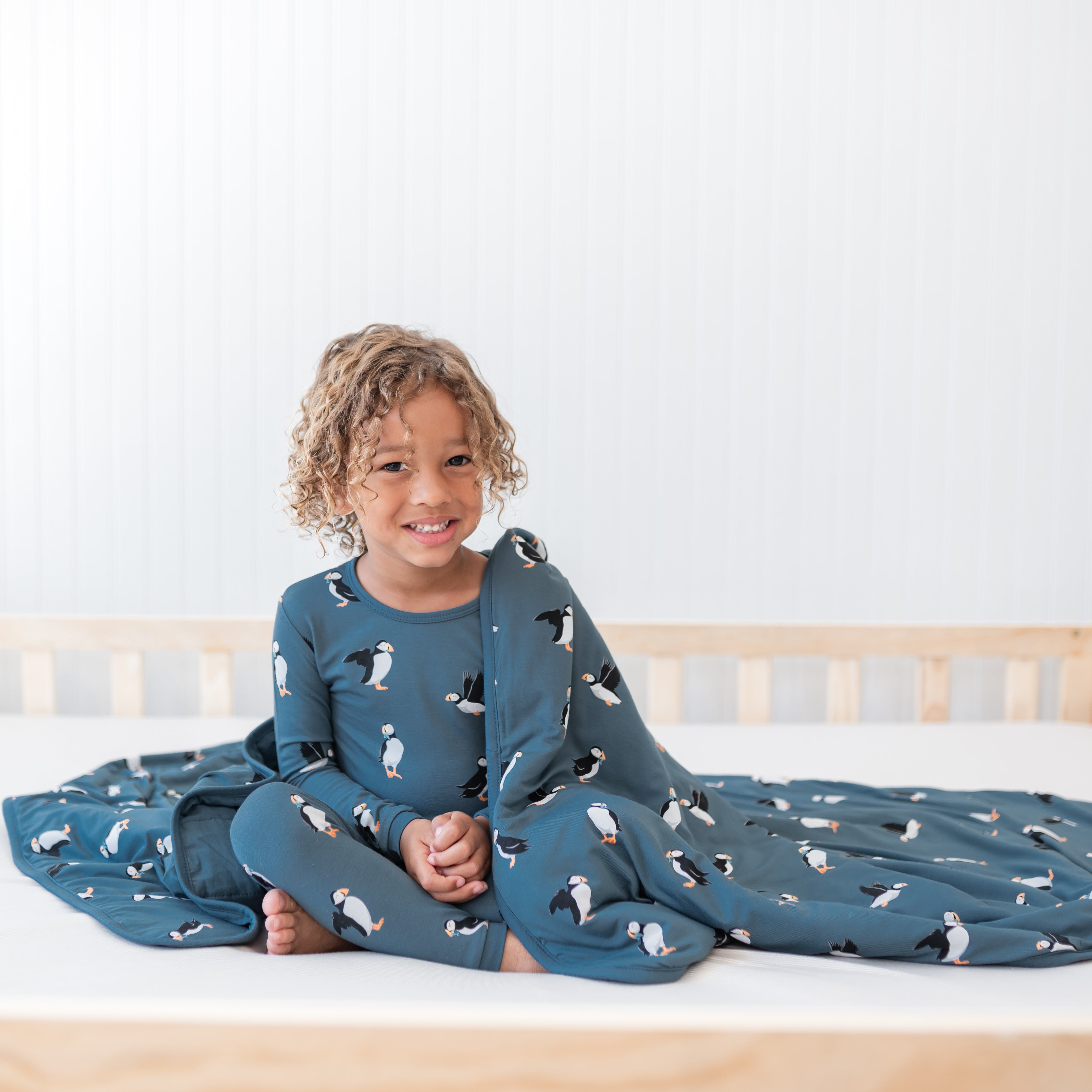 Young child sitting in a bed with the Toddler Blanket in Puffin 1.0 overtop of their shoulder and laying around them wearing matching long sleeve toddler pajamas