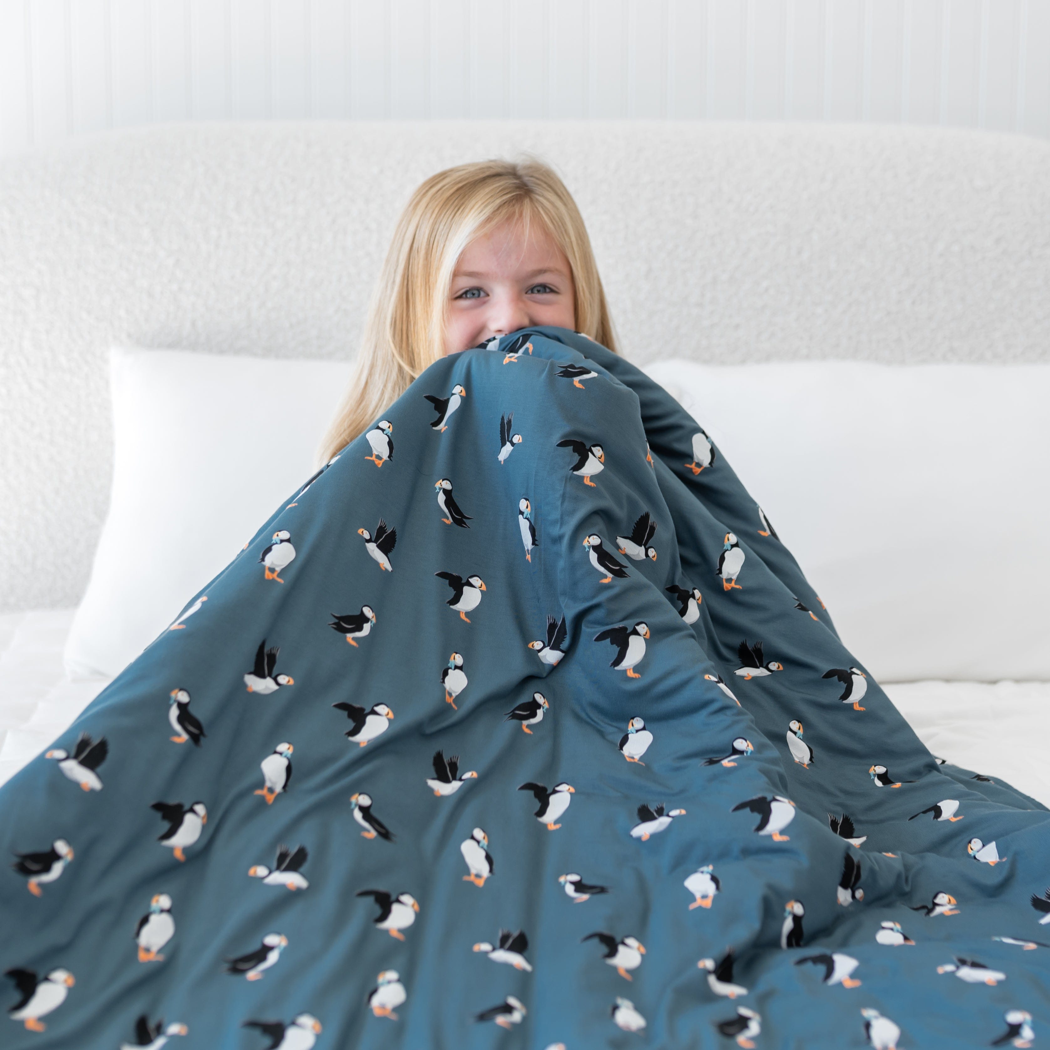 Young girl sitting on a bed holding the Youth Blanket in Puffin 2.5 up to her face
