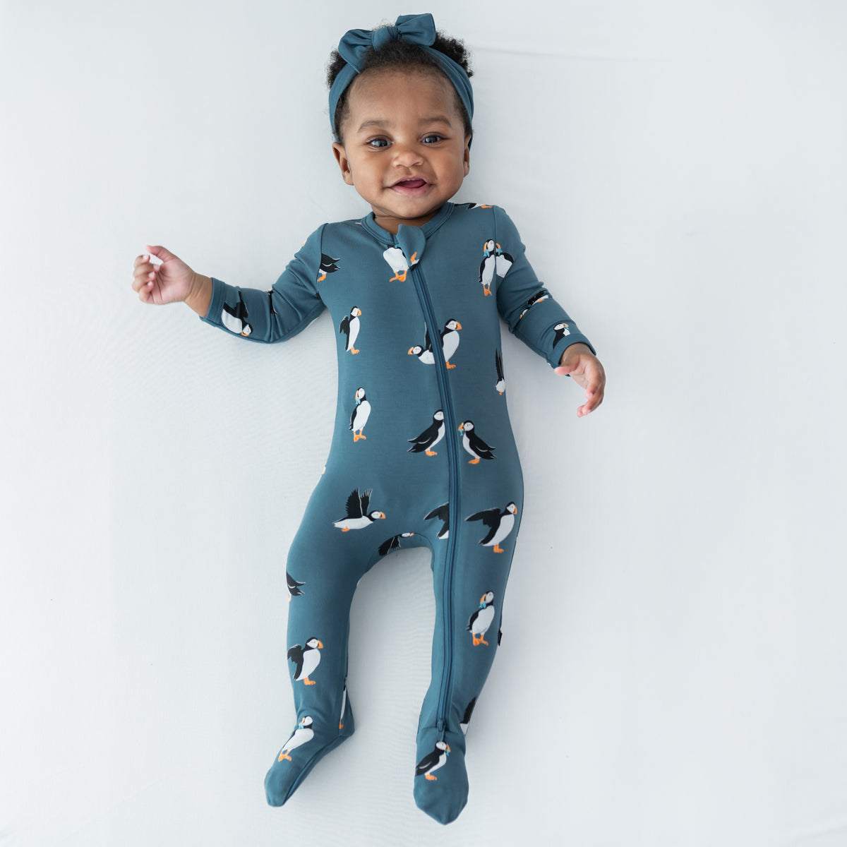Young infant laying on a light neutral surface wearing the Zippered Footie in Puffin with a knotted bow headband in atlantic