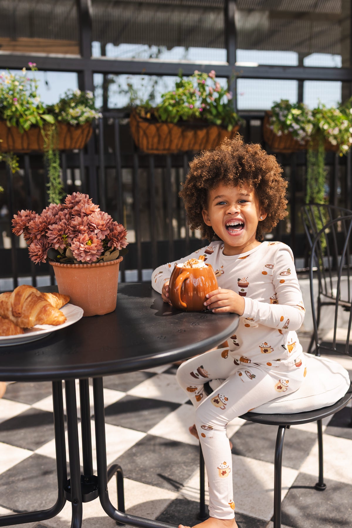 A young child sits smiling at an outdoor patio table, wearing a white long-sleeve pajama set with a brown coffee cup and mug pattern. The child is holding a small pumpkin decoration.