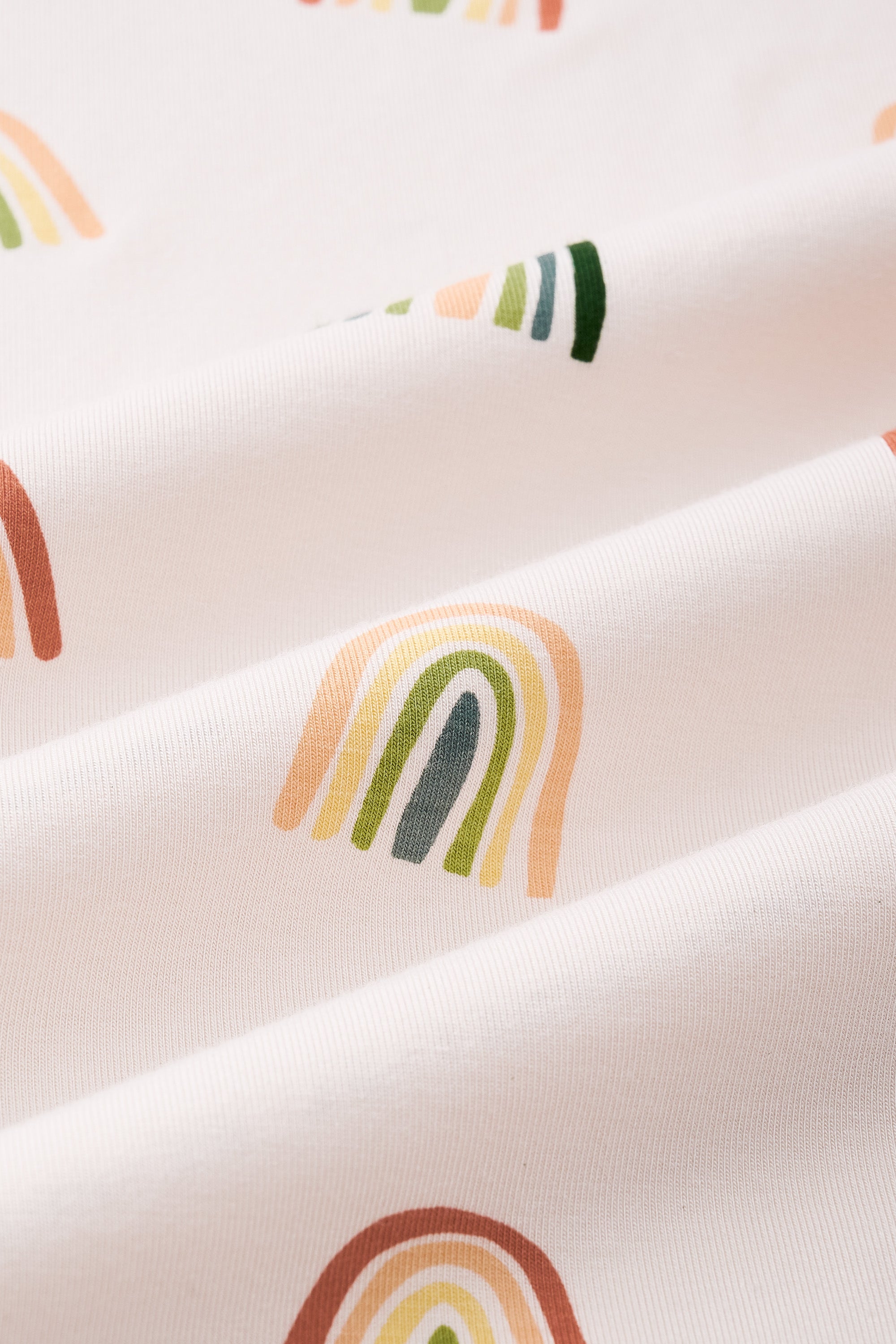 An all-over print featuring alternating rows of boho style rainbows in alternating directions over a light beige background color called Vanilla.
One row of rainbows has an outermost arc colored in a dark cool green, followed by a cool sage green, then a light olive green, and finished with an inner core colored in a light apricot.
Another row of of rainbows has an outermost arc colored in a burnt orange called Apricot, followed by a light peach, then a light yellow called Lilikoi, and finished with an
