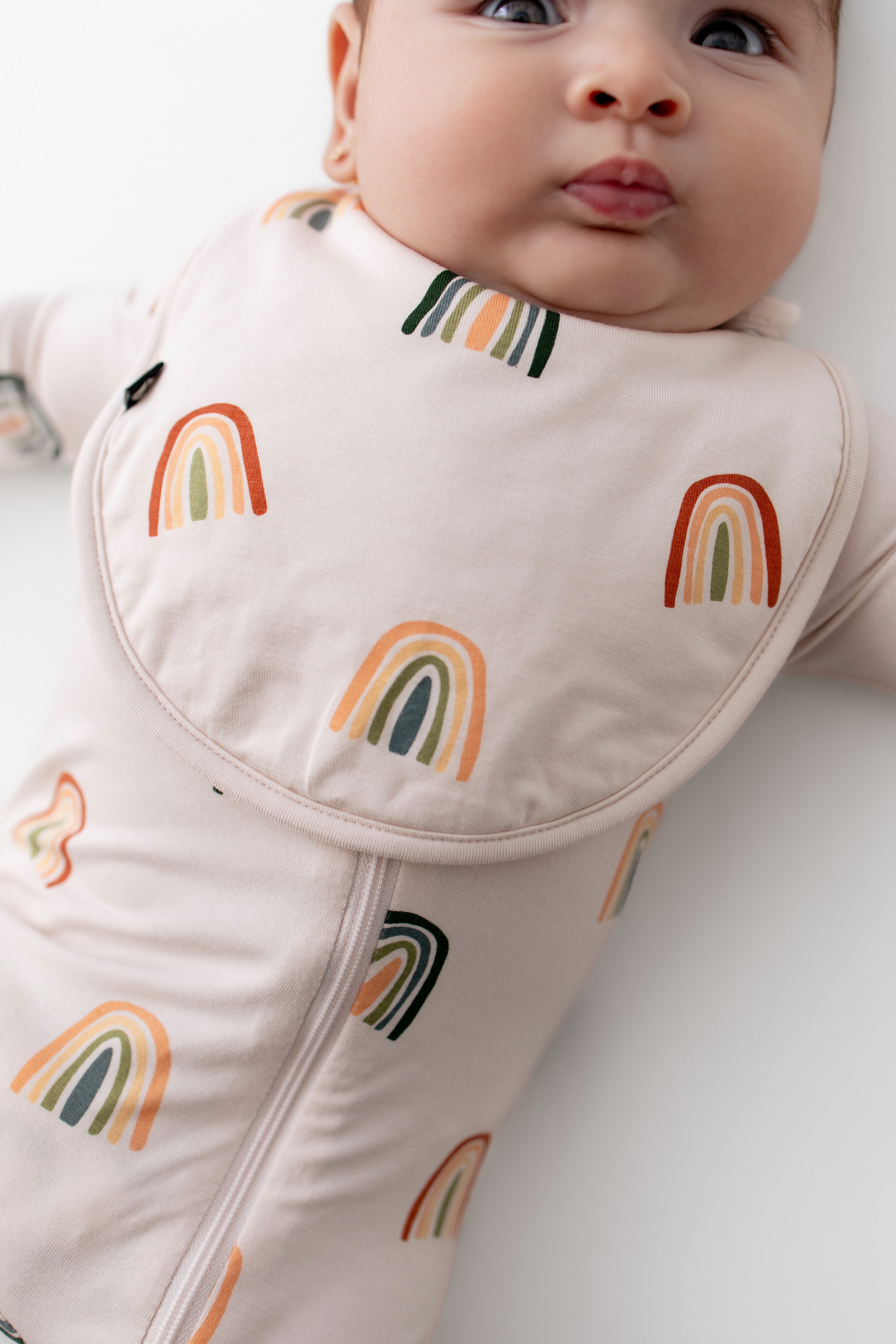 Baby wearing Cushy Bib in Retro Rainbow print over a similar print zippered romper on a white background