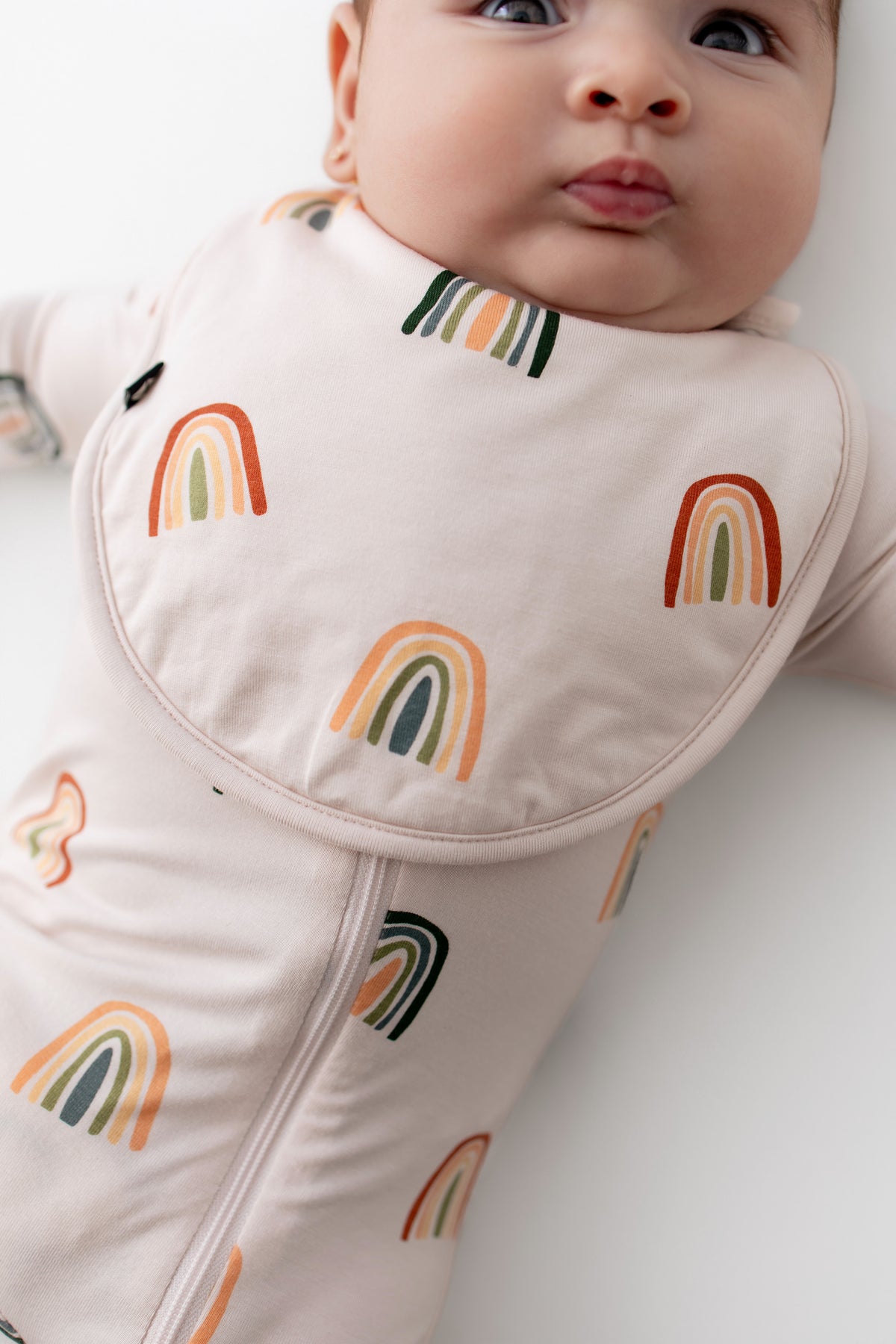 Baby wearing Cushy Bib in Retro Rainbow print over a similar print zippered romper on a white background