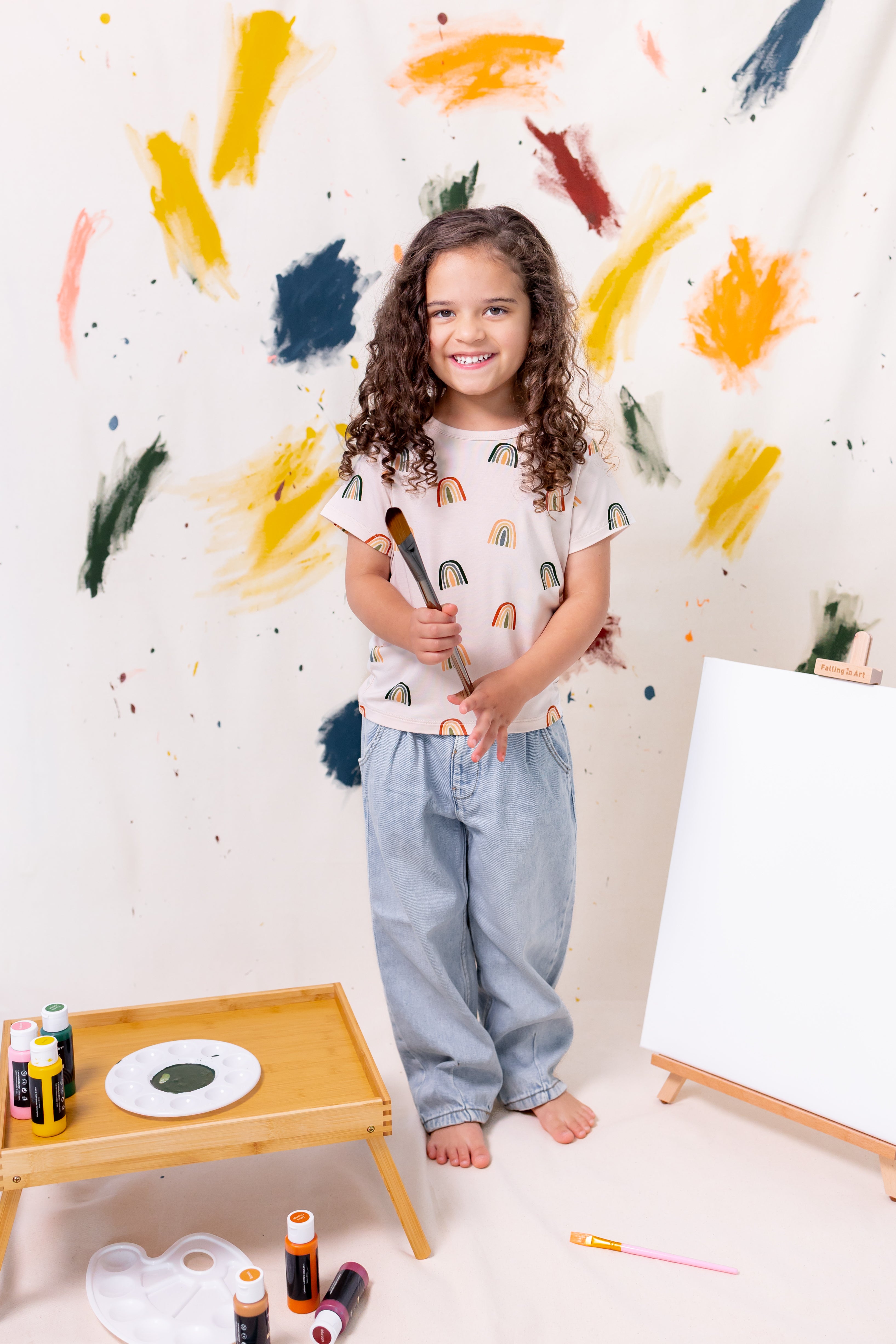 Girl posing in Toddler Basic Tee in Retro Rainbow print over blue pants holding a paint brush in front of a colorful background with few paints and canvas around her