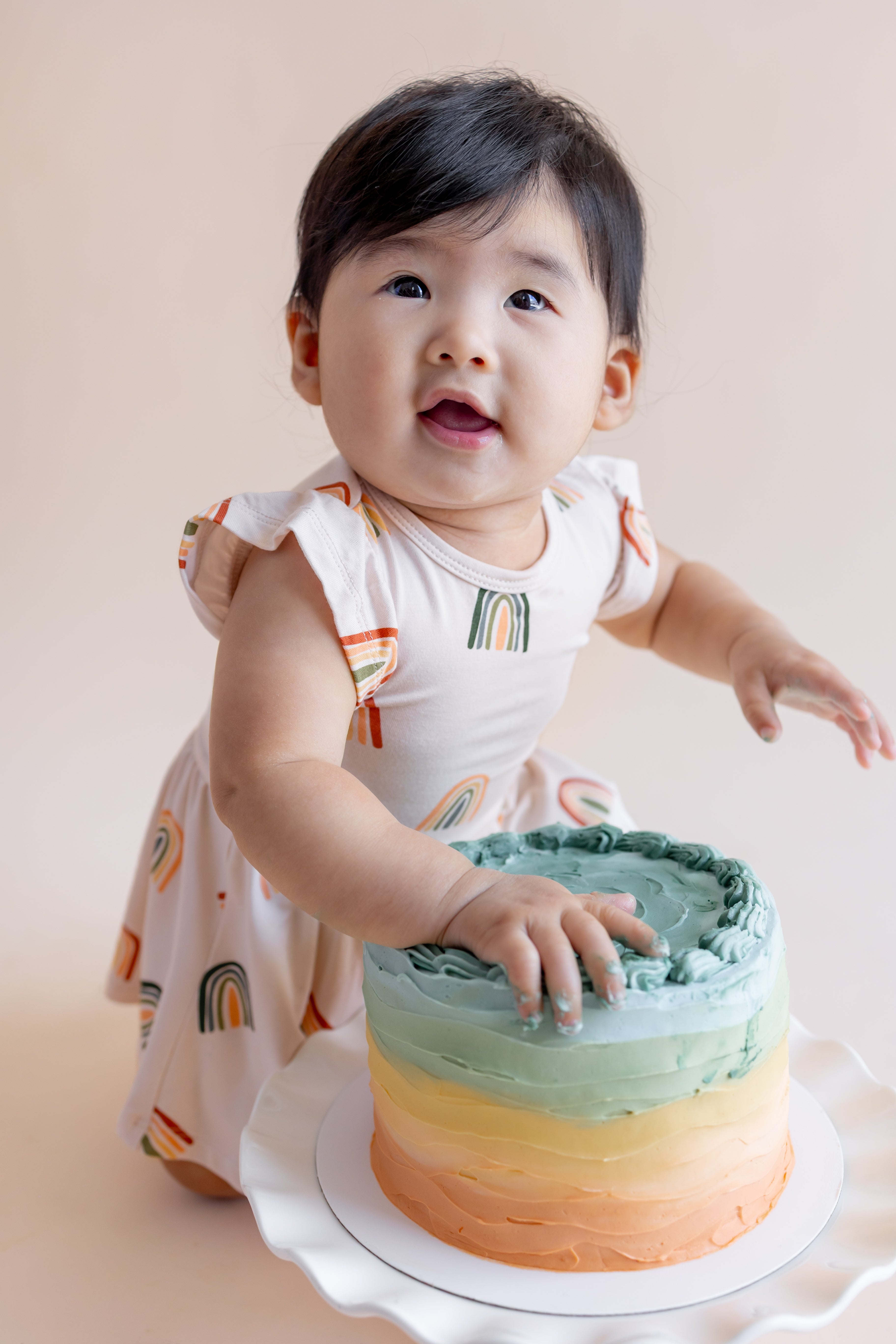 Baby wearing Twirl Bodysuit Dress in Retro Rainbow print sitting with a colorful cake on a beige background