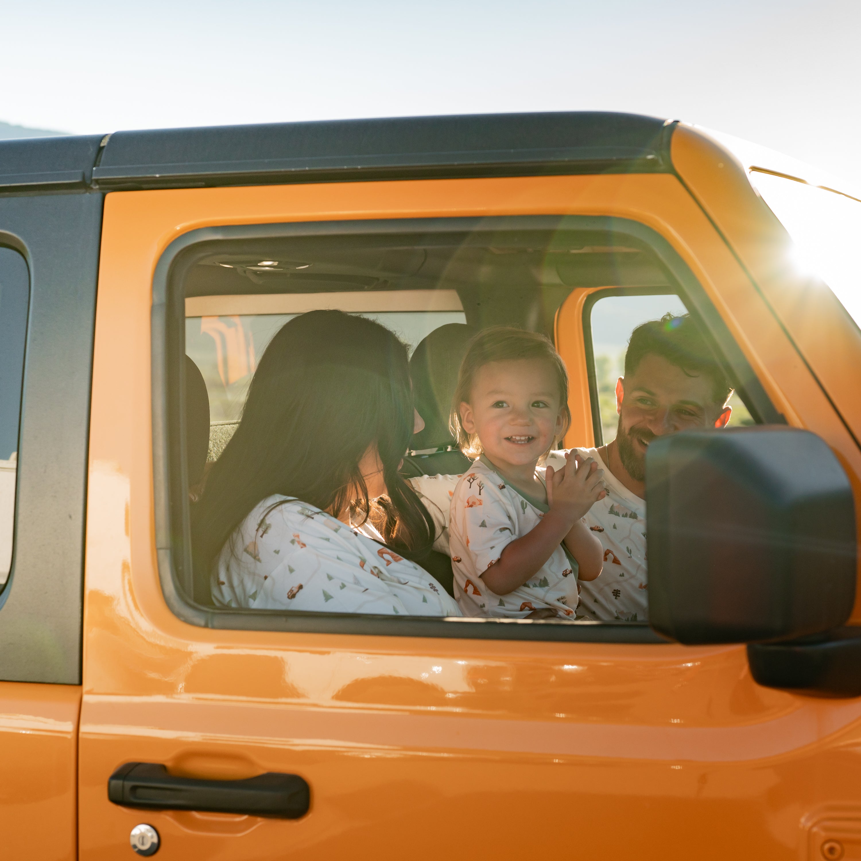 Family sitting in the front row of an Orange Jeep wearing shirts from the Road Trip collection. Pictured are the Women's Relaxed Fit V-Neck, Toddler Crew Neck Tee and Men's Crew Neck Tee