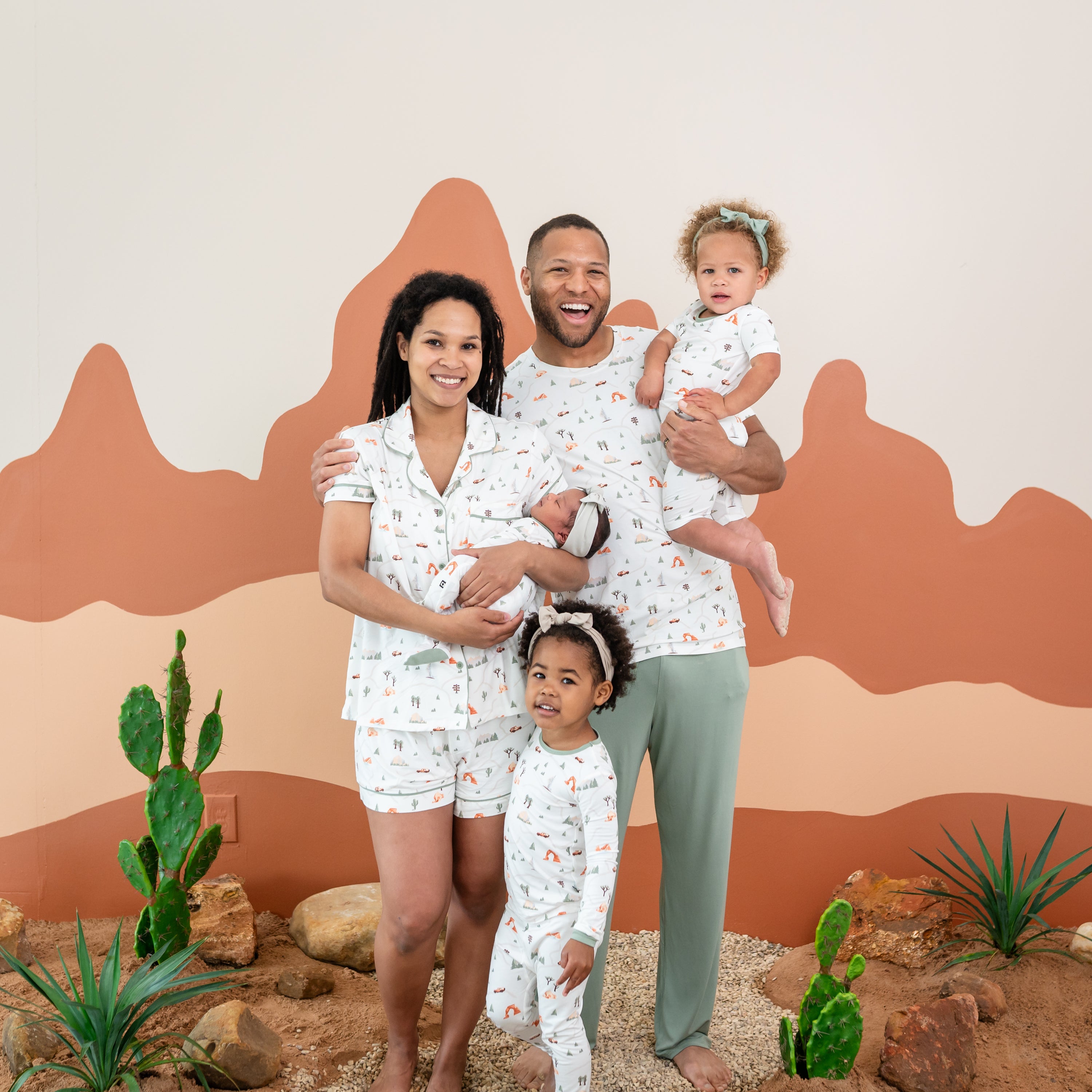 Family of five wearing the Road Trip Collection. Mother wearing the Short Sleeve Women's Pajama Set in Road Trip holding her newborn in a matching footie. Father wearing the Men's Crew Neck holding his daughter who is matching in a Short Sleeve Pajama set in Road Trip. Oldest daughter standing in front of mother and father wearing the Long Sleeve Pajama Set in Road trip