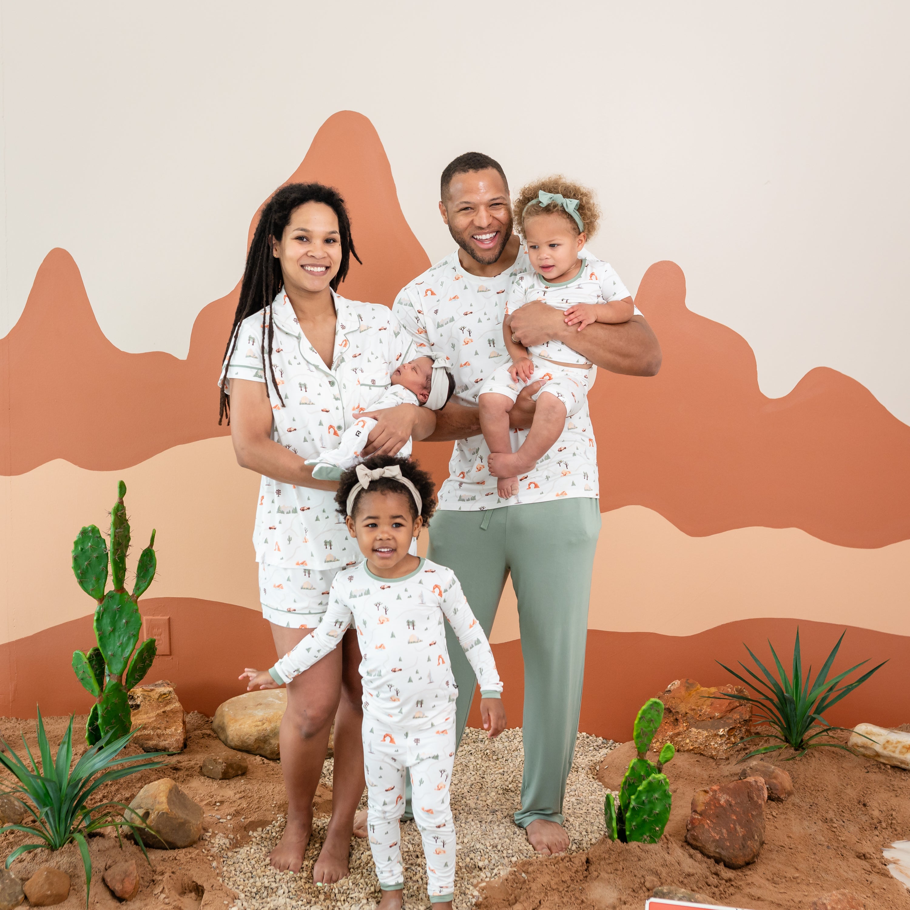 Family of five wearing the Road Trip Collection. Mother is wearing the Women's short sleeve pajama set holding her newborn who is in a footie. Father is wearing a Men's Crew Neck holding his daughter wearing the Short Sleeve toddler pajamas. The oldest daughter is standing in front of parents wearing the long sleeve toddler pajamas.