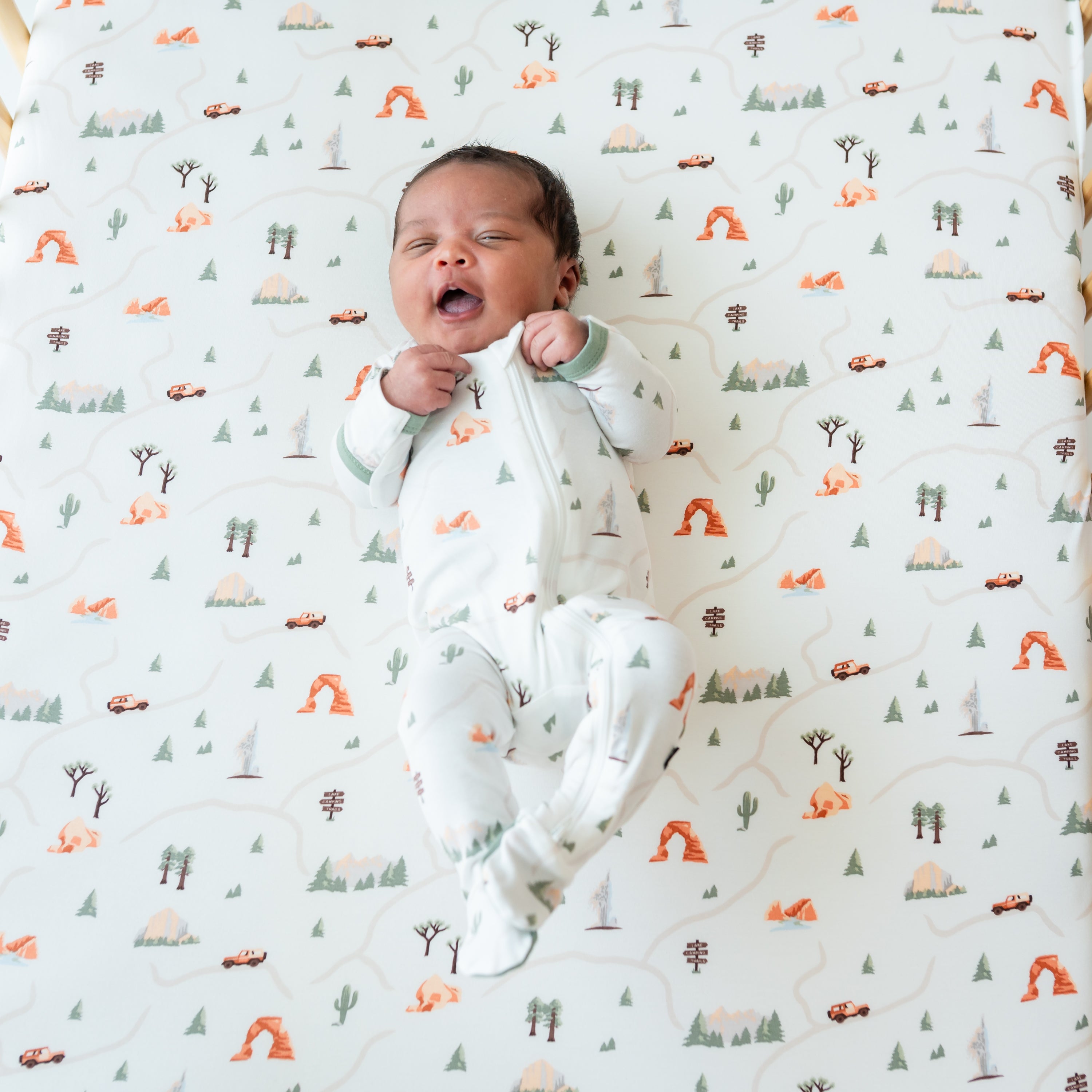 Newborn wearing Zippered Footie in Road Trip laying on a matching crib sheet for camping themed nursery