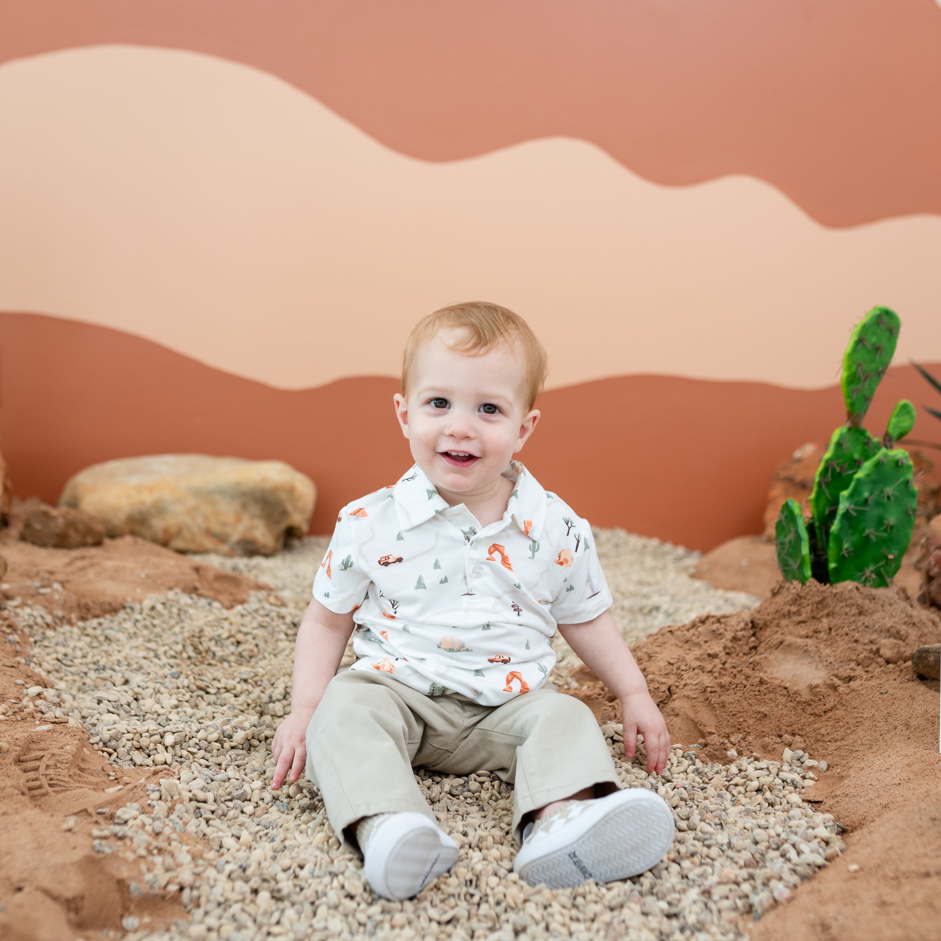 Toddler model sitting in rocks wearing the Toddler Short Sleeve Polo in Road Trip with a desert inspired back drop