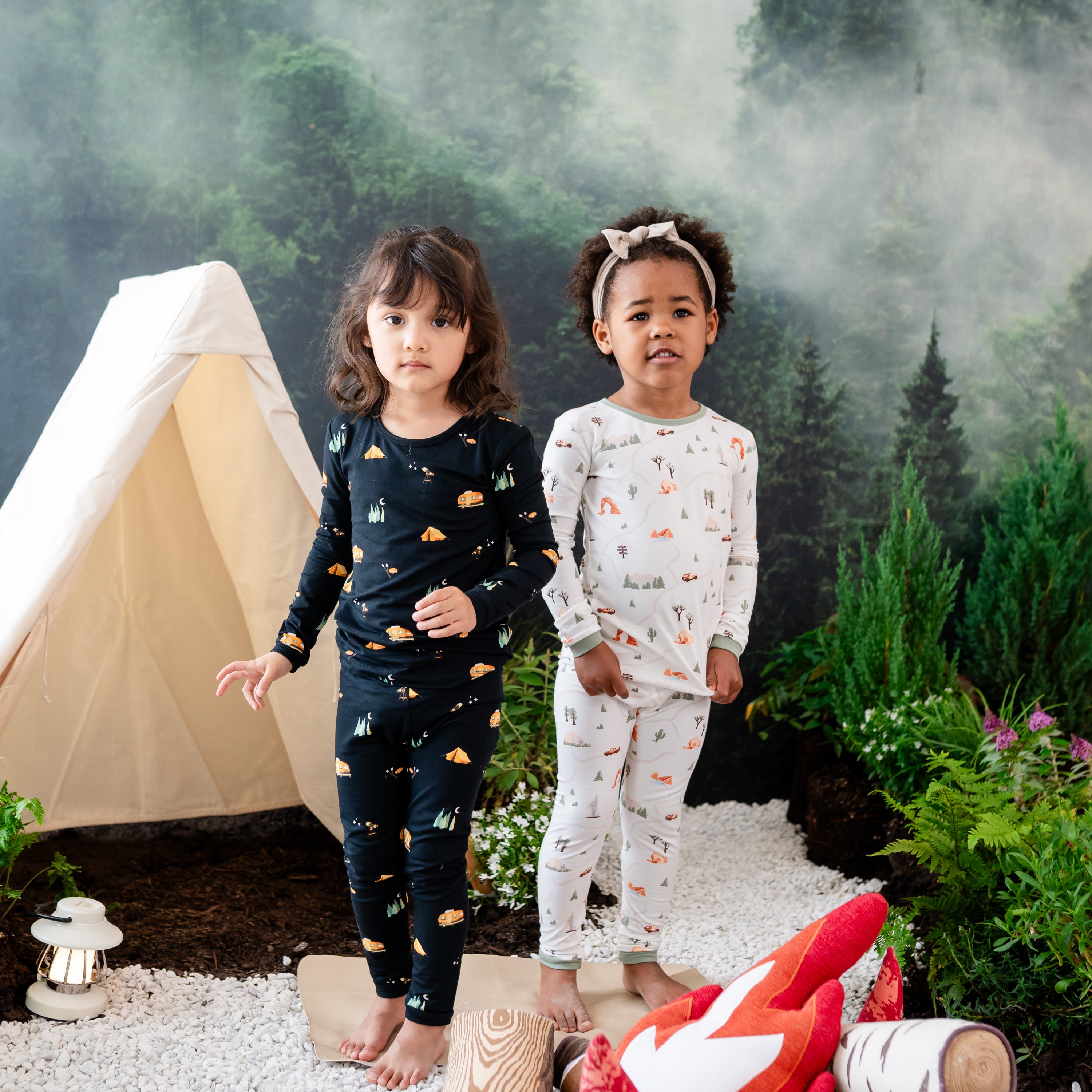 Two models standing side by side wearing the long sleeve toddler pajama set in Under the Stars and Road Trip in front of a camping set up