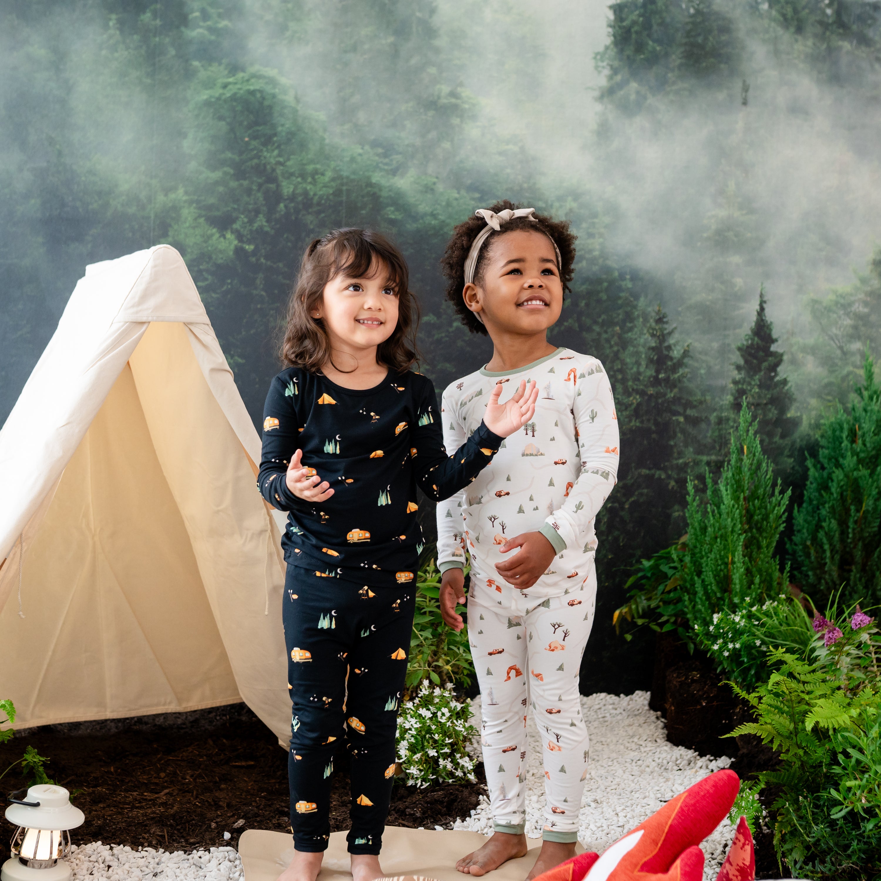 Two girl models wearing long sleeve toddler pajamas in Under the Stars and Road Trip standing in front of a camping back drop