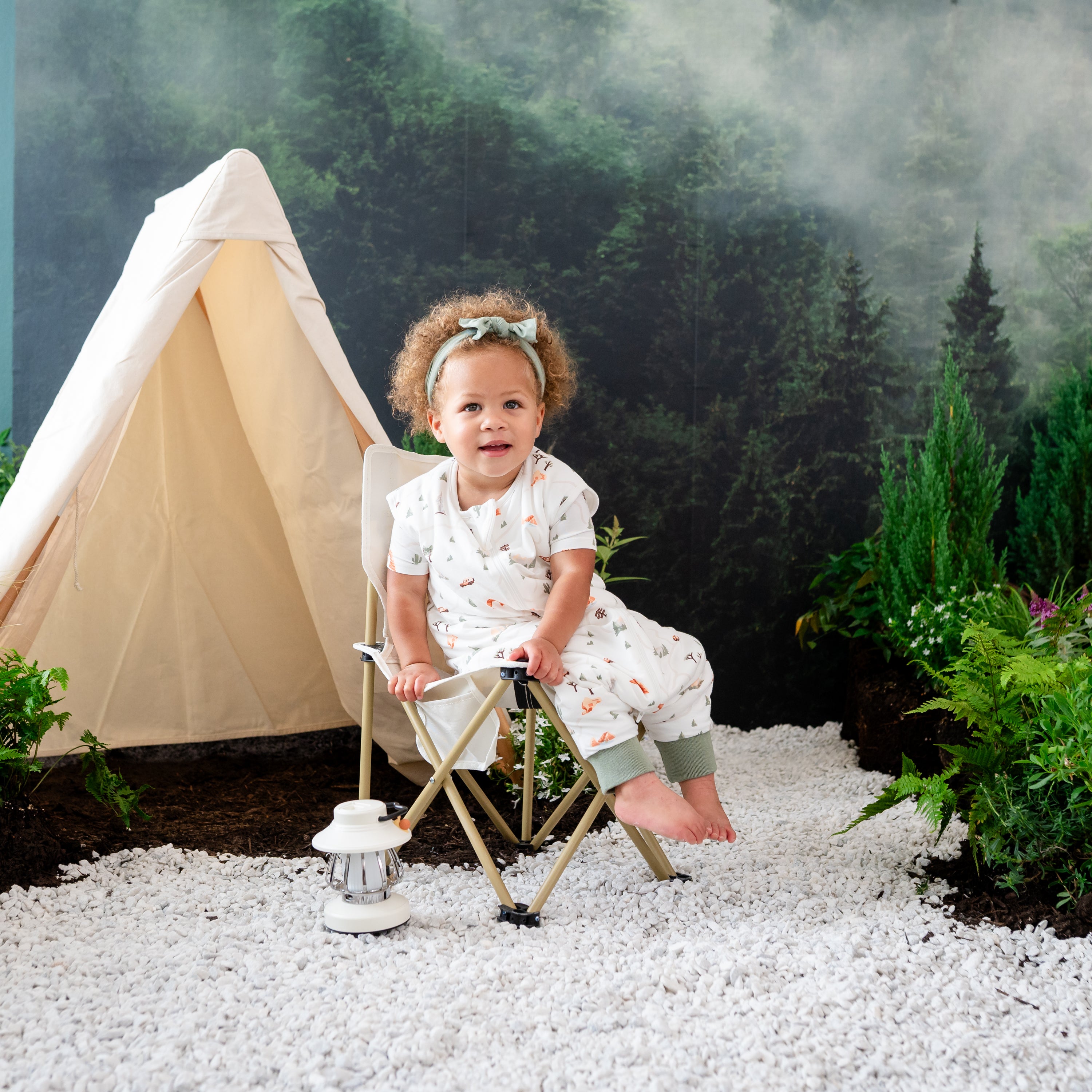 Child model sitting in a canvas lawn chair with a wilderness inspired camping backdrop wearing the Sleep Bag Walker in Road Trip 1.0