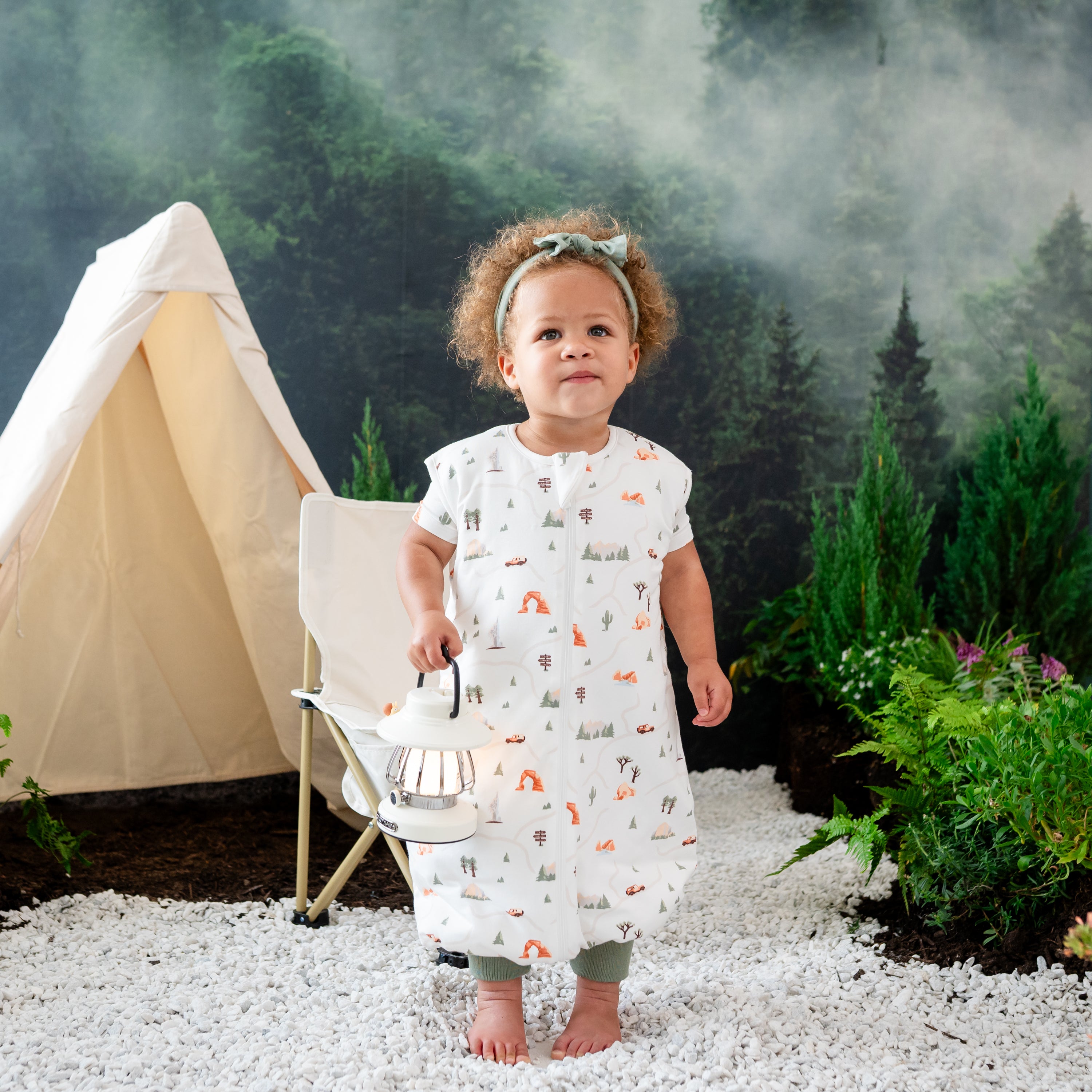 Toddler girl standing in rocks holding a light lantern wearing the Sleep Bag Walker in Road Trip 1.0 with a wilderness camping backdrop