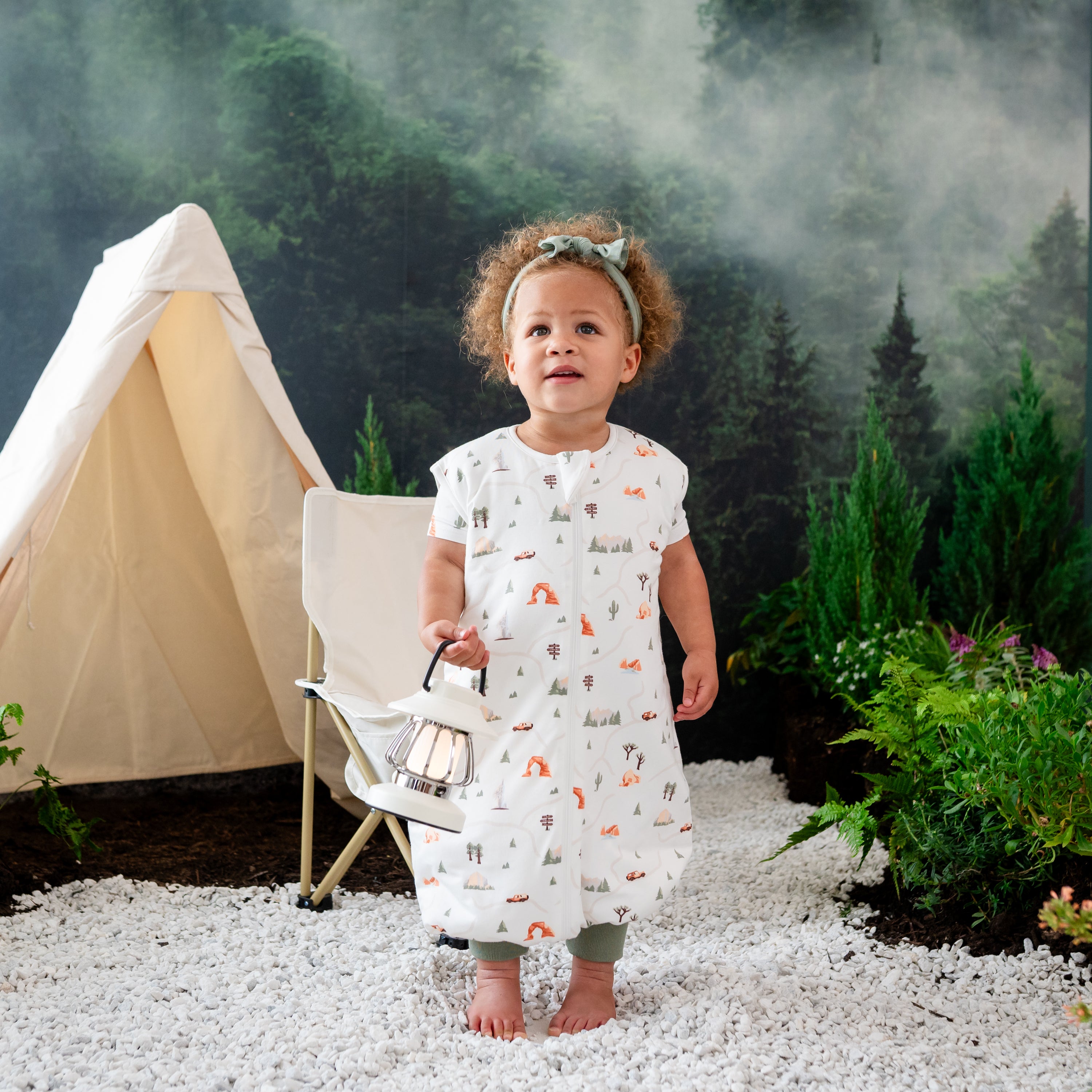 Toddler girl standing in rocks in front of a camping wilderness inspired backdrop wearing the Sleep Bag Walker in Road Trip 1.0